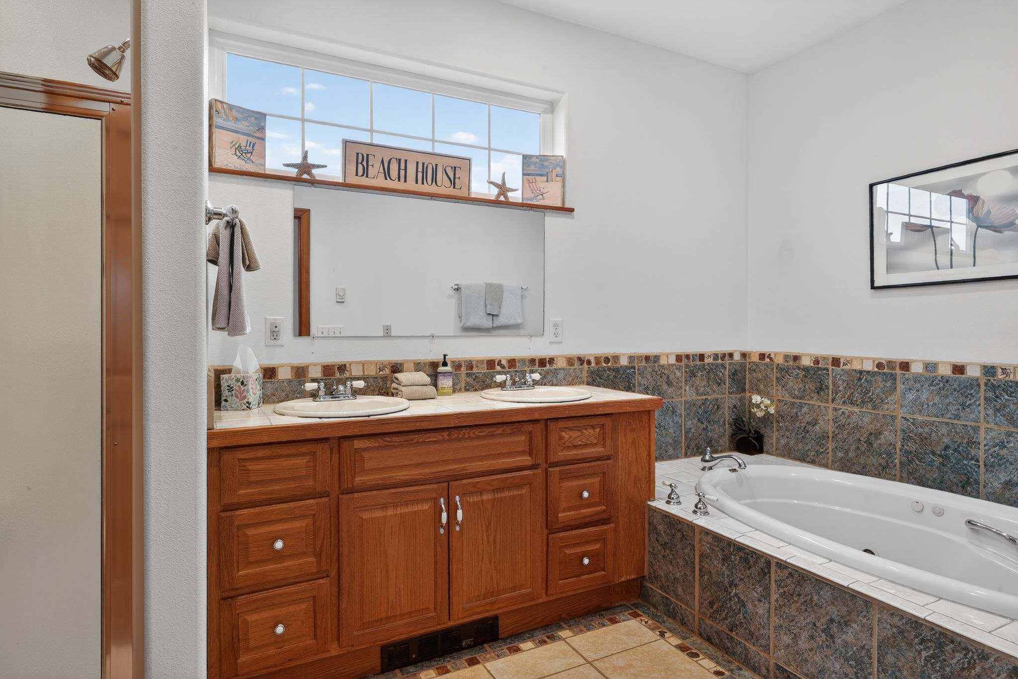 Bright bathroom with a large window, tiled floors, and a double vanity with a wood cabinet and tiled backsplash, next to a large soaking tub with chrome fixtures.