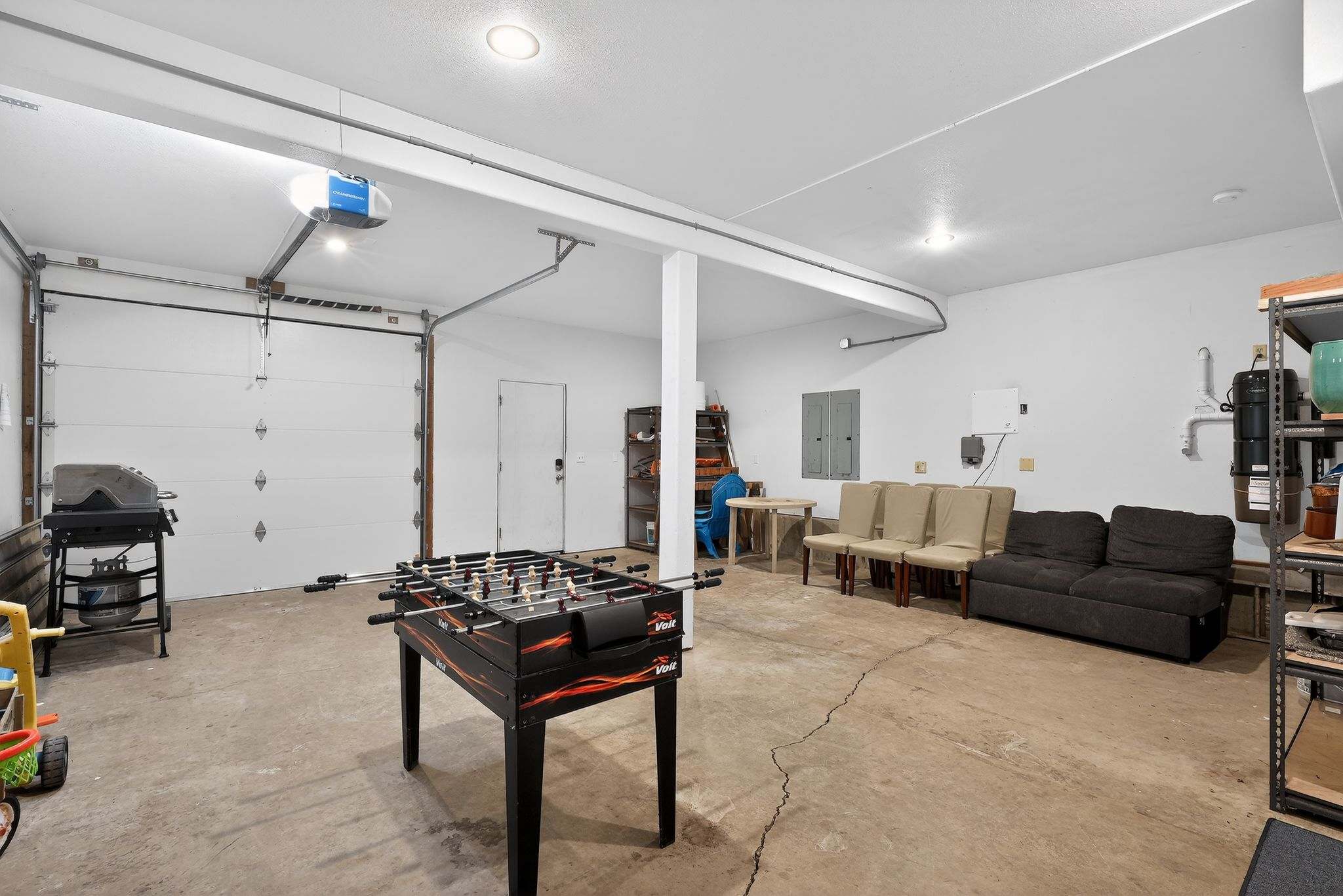 A spacious garage has been converted into a recreational area, featuring a foosball table in the foreground and a casual seating arrangement with chairs and a sofa in the background. A large garage door and shelving units are also visible.