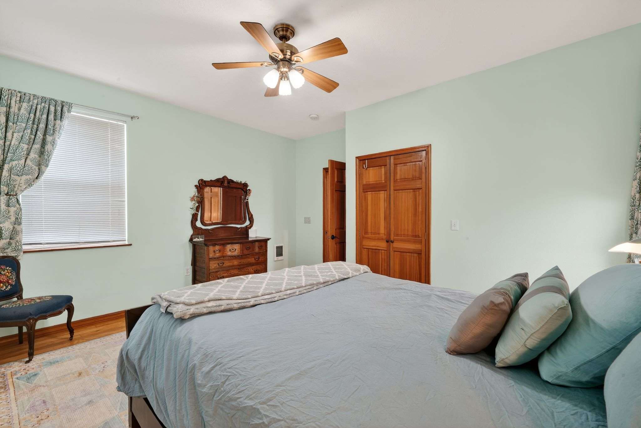 A bedroom with light mint green walls and hardwood floors features a dark wooden bed with light blue bedding. A wooden sliding closet is on the back wall, and an open doorway leads to another area.