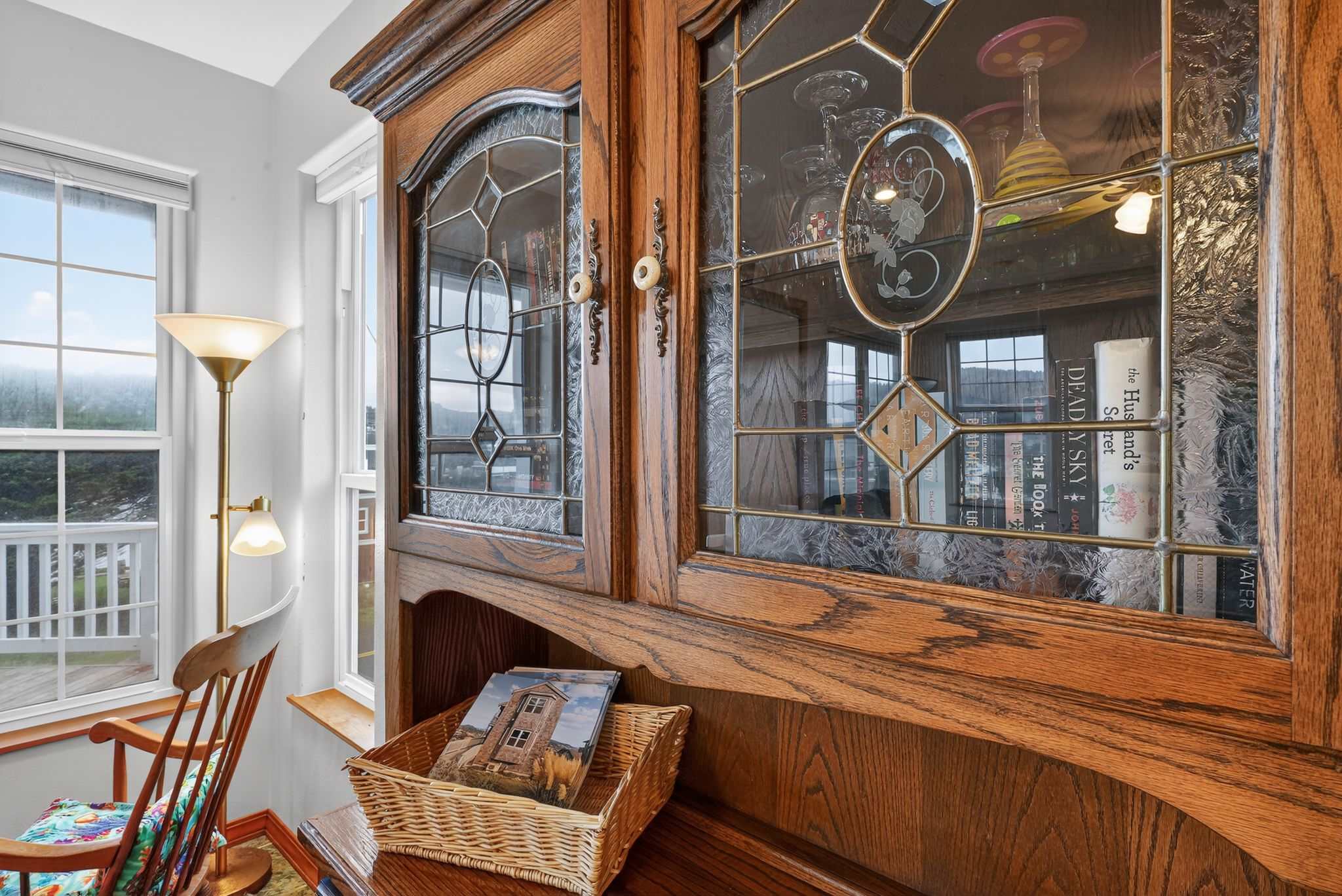 A detailed shot highlights an ornate wooden cabinet with leaded glass doors, showcasing decorative items inside and a woven basket on a lower shelf. A window to the left offers a glimpse of an outdoor deck and distant views.