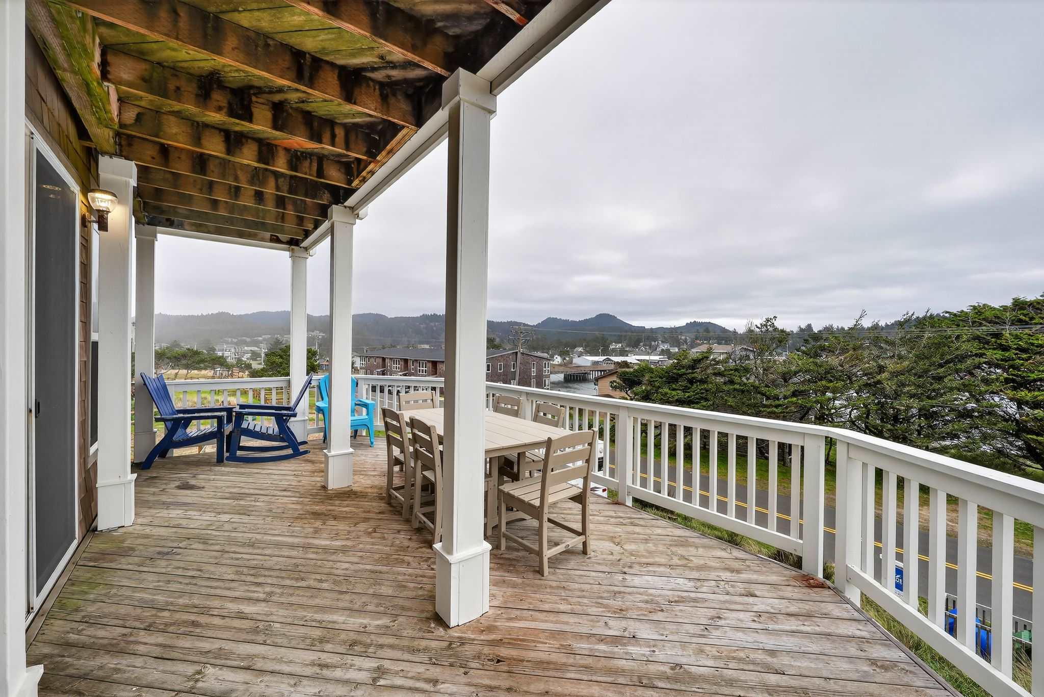 A spacious wooden deck features a light wood dining table with six chairs and two blue Adirondack chairs, all enclosed by white railings. The deck overlooks a town, distant hills, and a road under a cloudy sky.