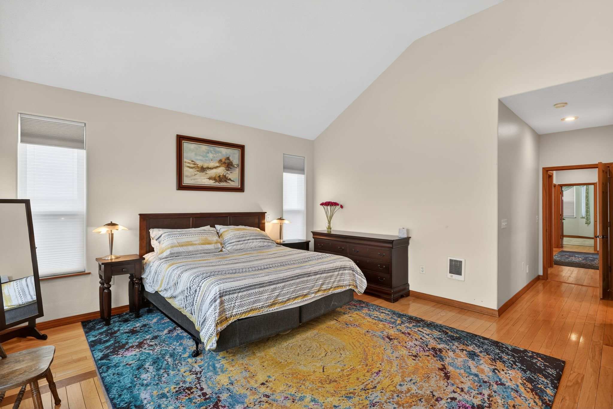Spacious bedroom with a vaulted ceiling and light walls, featuring a large bed with striped bedding, two nightstands with lamps, a dark wood dresser, and a colorful abstract rug on hardwood floors.