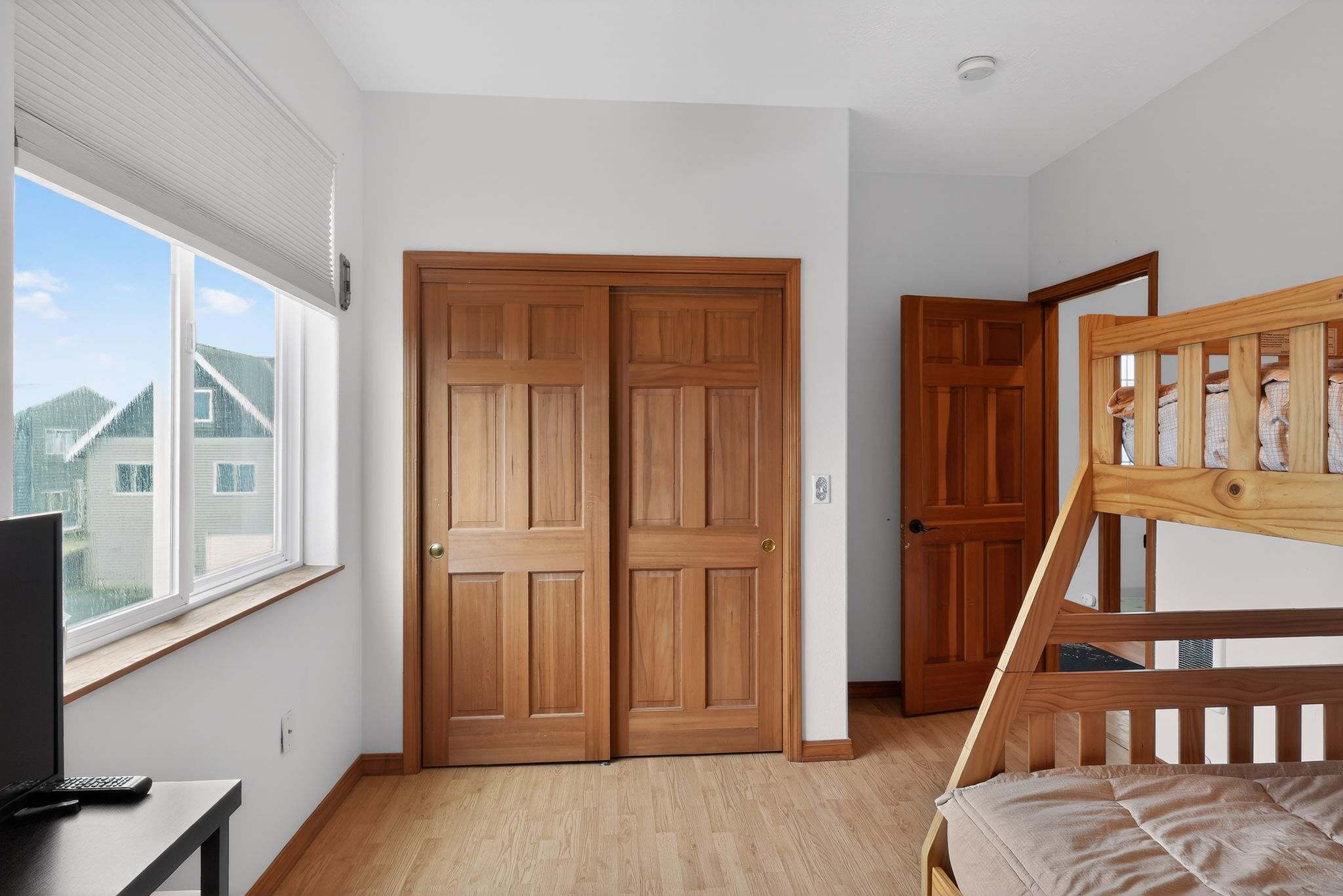 A bright bedroom with light wood flooring features a wooden bunk bed on the right and a large window with white blinds on the left, offering a view of neighboring houses. A two-door wooden sliding closet is centered on the back wall.