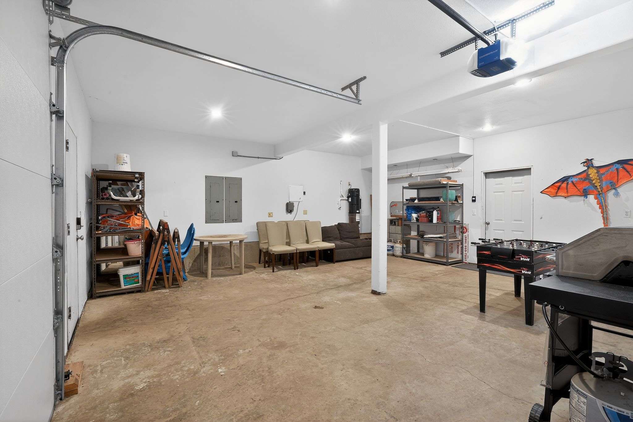 A spacious garage with a concrete floor, white walls, and exposed ceiling beams, featuring a foosball table, seating, and storage shelves, illuminated by bright overhead lights.