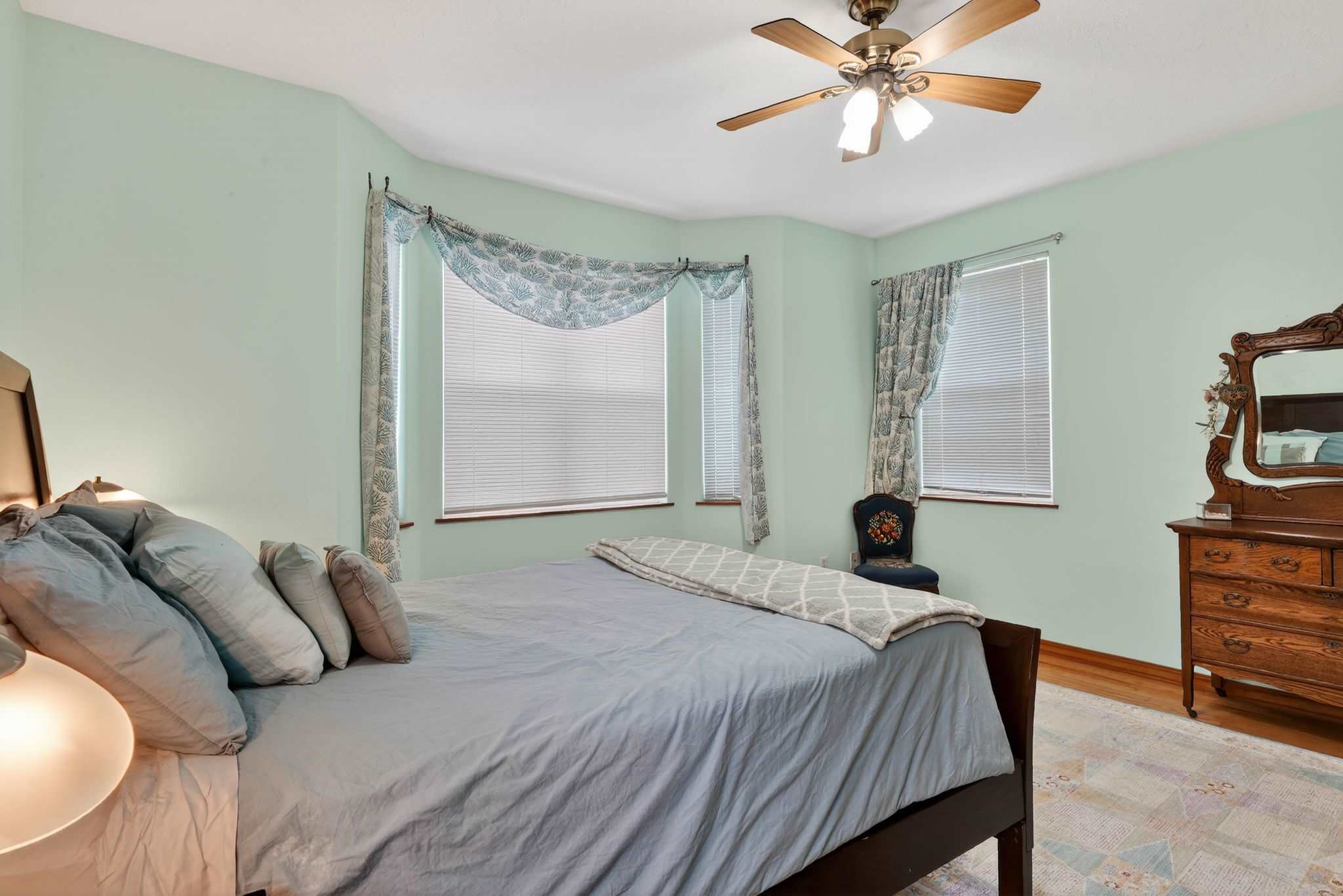 A bedroom with light mint green walls and hardwood floors features a dark wooden bed with light blue bedding. A bay window with blinds and patterned valances provides natural light, and a wooden dresser with a mirror is visible.