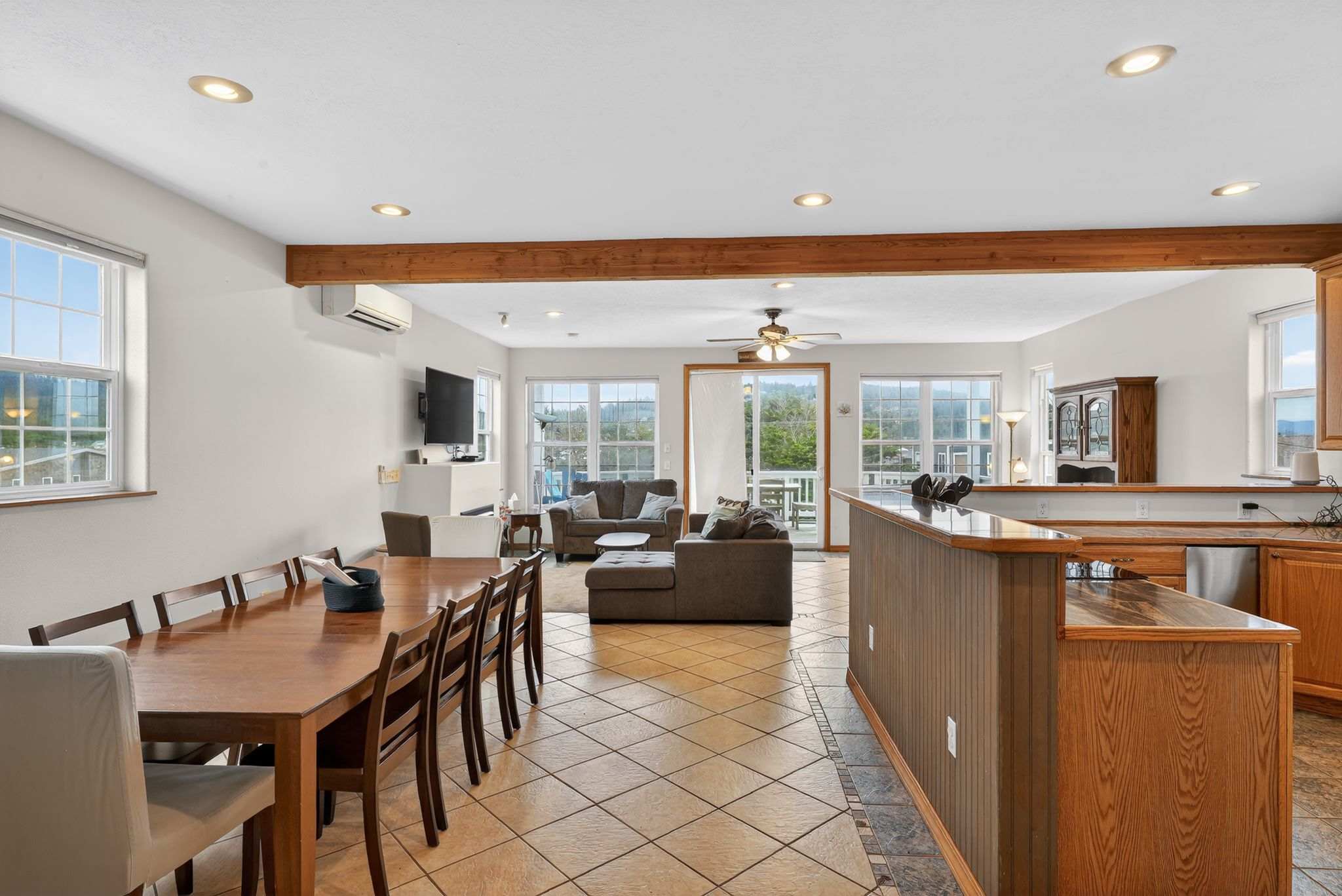A long wooden dining table with multiple chairs dominates the foreground, part of an open-concept space that flows into the living room and kitchen. Tile flooring unifies the area, with a large wooden beam overhead and windows offering views.
