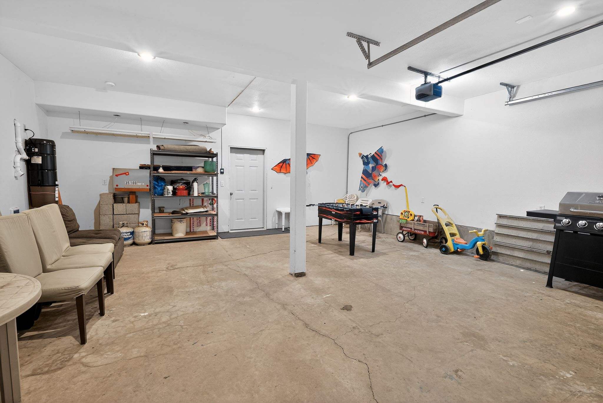 A wide view of a bright garage space with a concrete floor, white walls, and a foosball table, alongside storage shelves, a grill, and children's toys, suggesting a multi-functional recreation area.