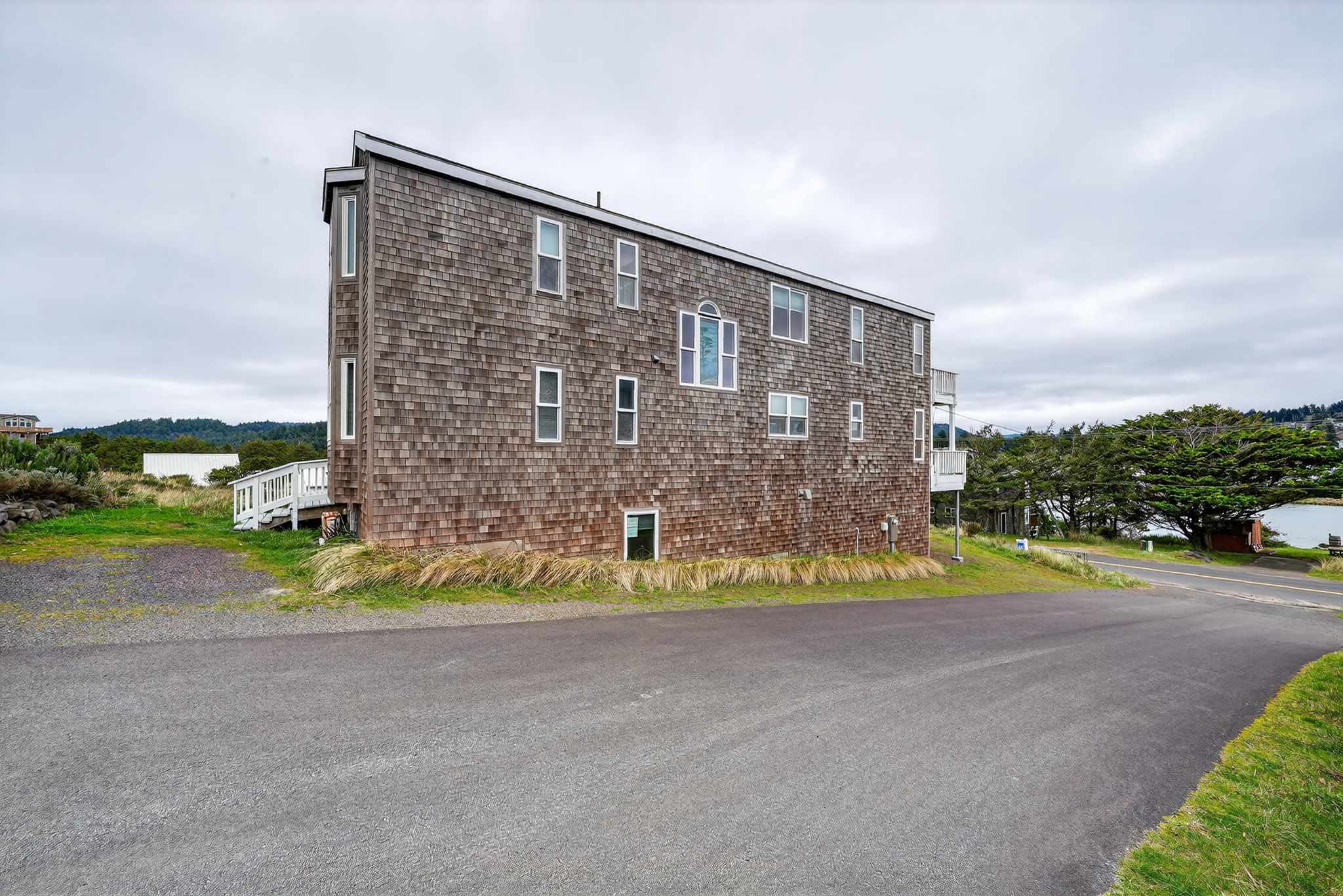 The long, narrow cedar shake house is viewed from the side, showcasing its multiple windows and the paved road alongside it. The property is situated on a slight incline with grassy areas.