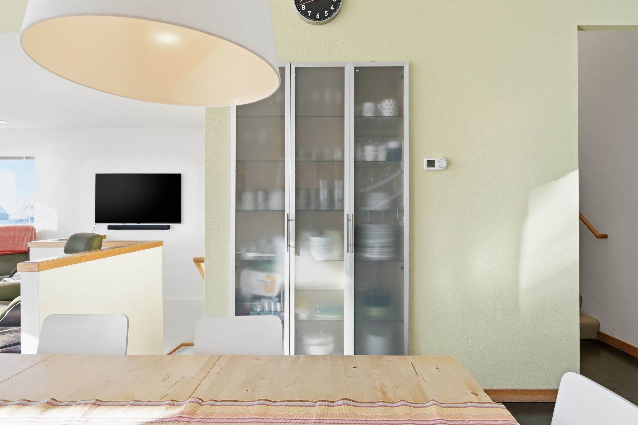 Close-up of a tall, modern cabinet with frosted glass doors, revealing dishes and kitchenware inside. A wall clock is mounted above, and light green walls frame the area.
