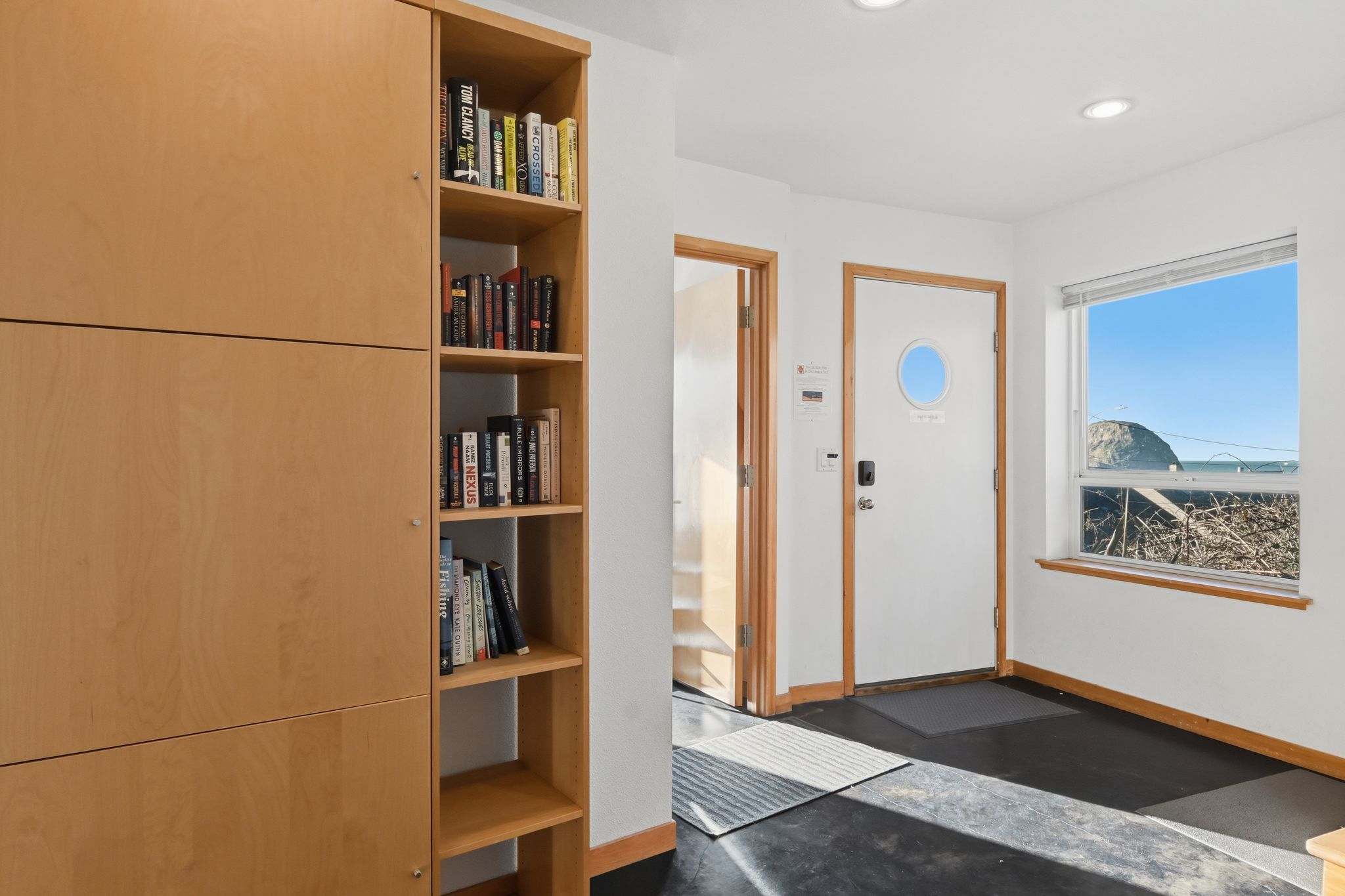 A bright entryway features a built-in wooden bookshelf filled with books, a dark floor, and white walls. A white door with a porthole window and a keyless entry system is visible, along with a large window offering a glimpse of the ocean.
