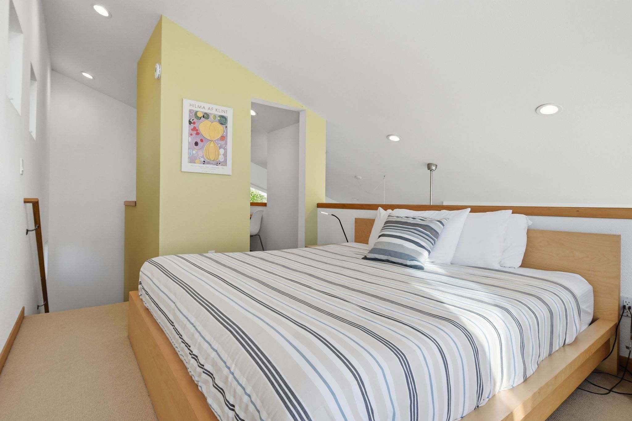 A bright bedroom with a light wood bed frame, striped bedding, and a light yellow accent wall. A modern art print hangs on the wall, and recessed lighting illuminates the sloped ceiling.