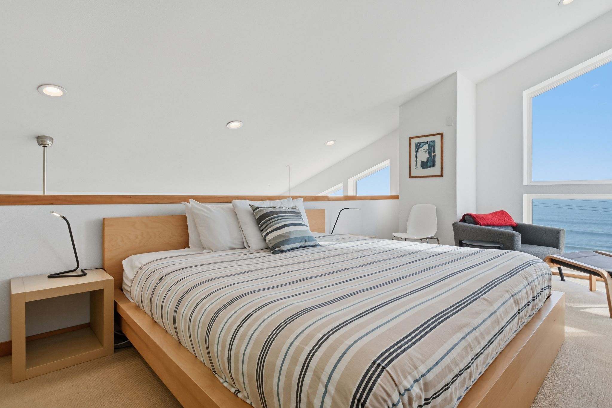 Bright bedroom featuring a king-sized bed with striped bedding and light wood frame. Large windows offer stunning ocean views, and a modern armchair sits in the corner.