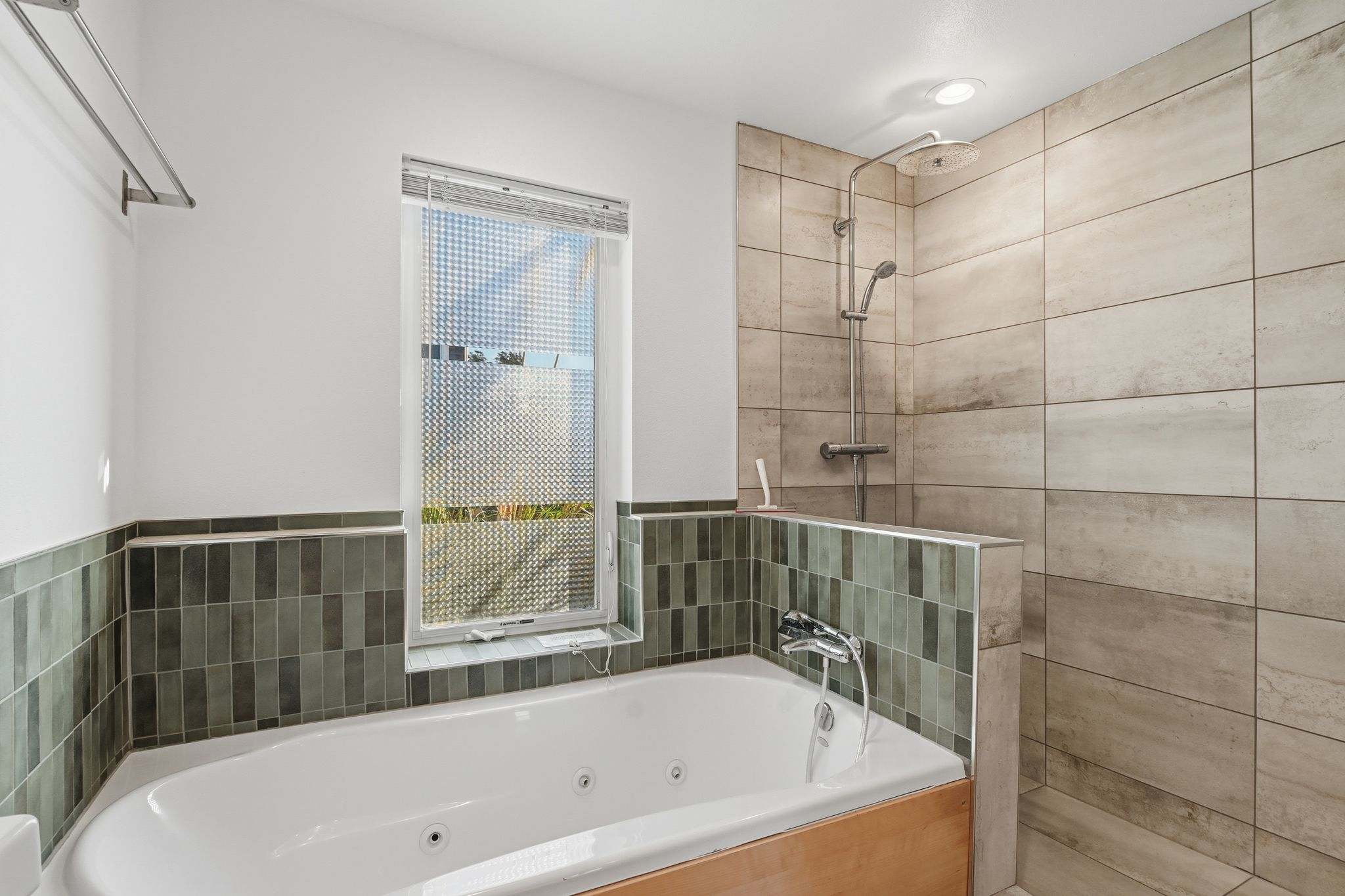 A close-up view of a bathroom's jetted bathtub with a wooden surround and an adjacent shower area. The walls are tiled in light grey and green, and a window with privacy blinds is above the tub.