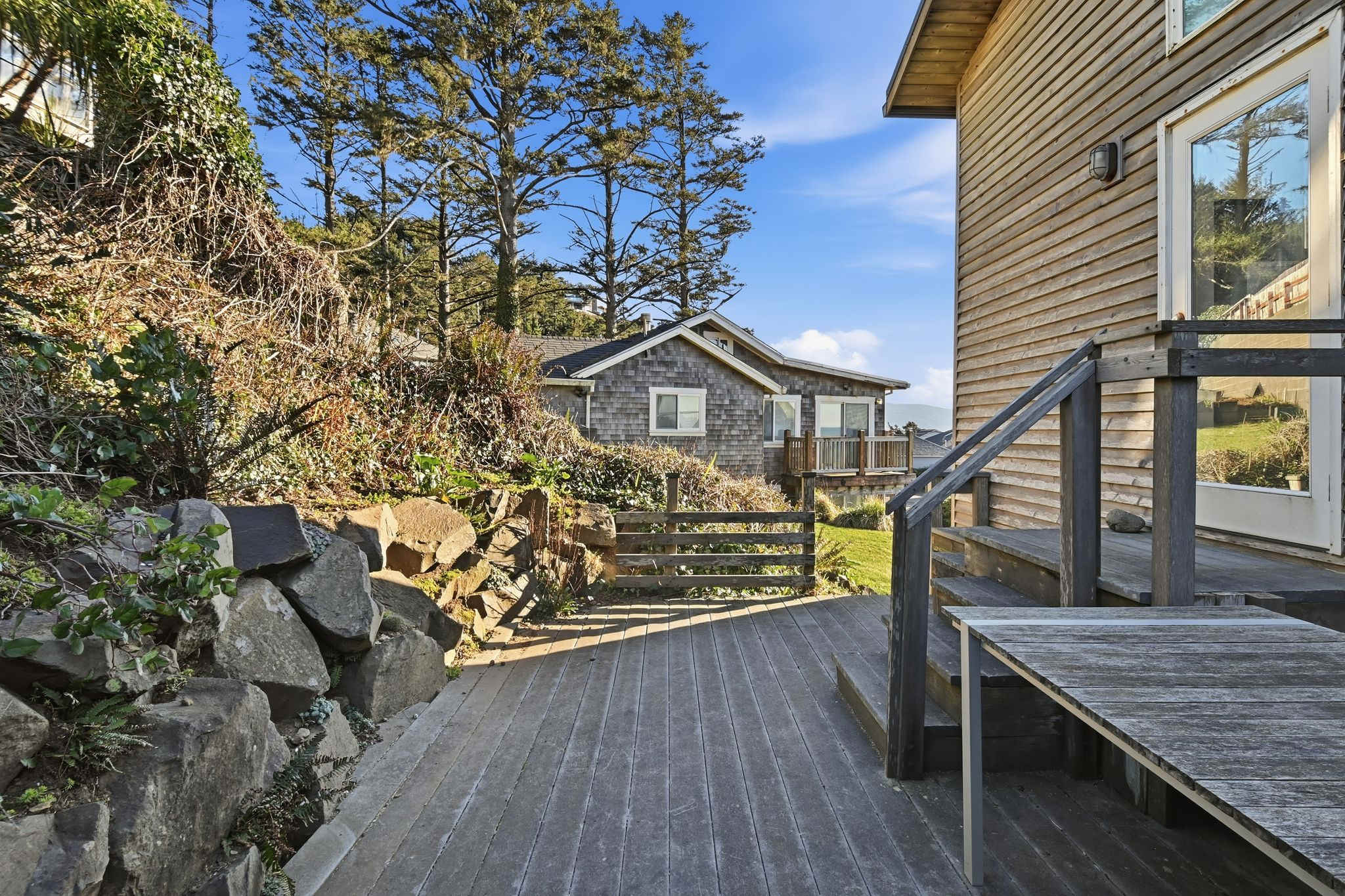 A wooden deck with steps and a railing is surrounded by natural landscaping, including a rock wall and various shrubs. Tall evergreen trees and a glimpse of another shingle-sided house are visible in the background under a clear blue sky.