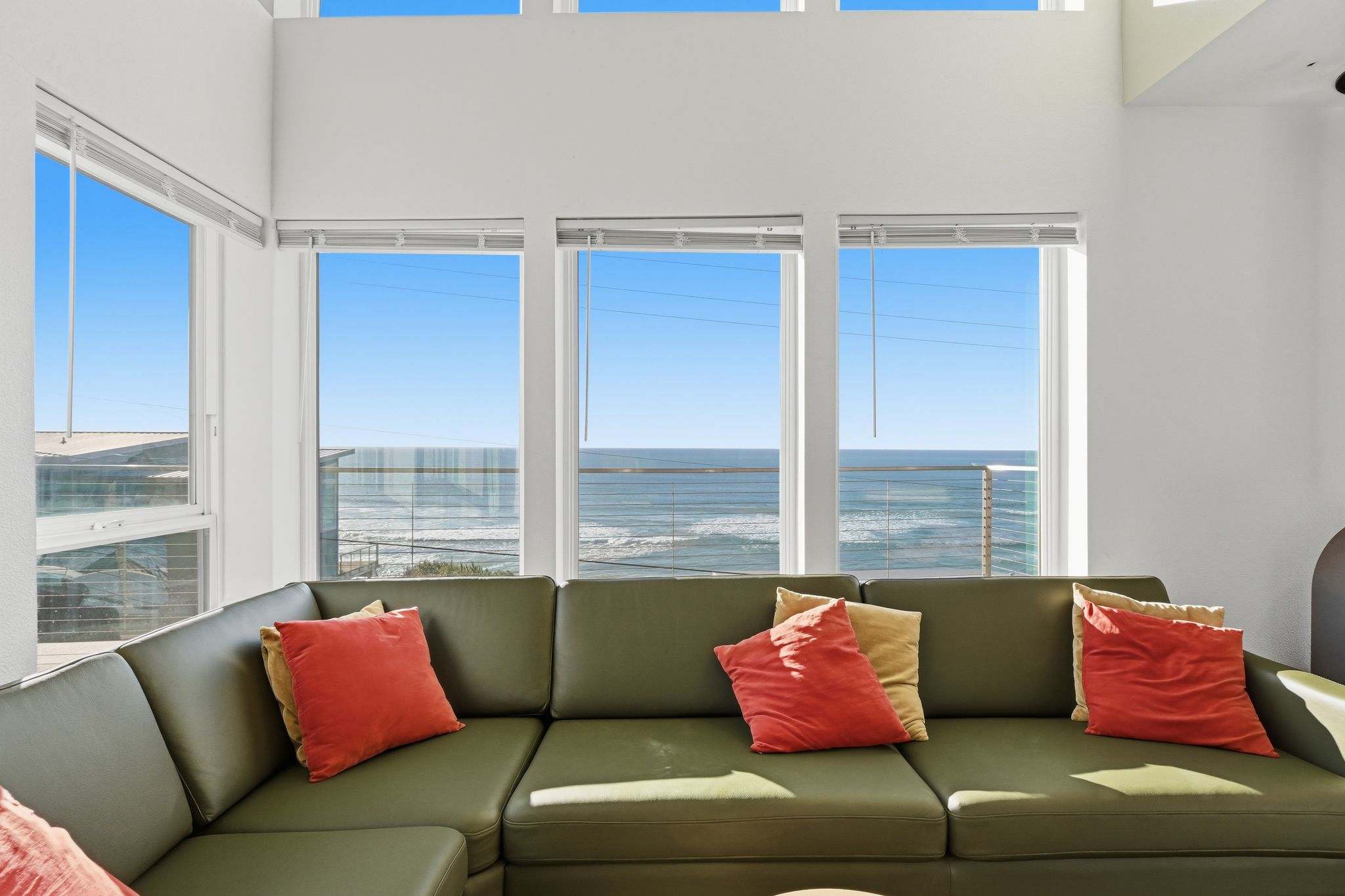 A spacious living room features a large olive green sectional sofa adorned with red and yellow throw pillows, facing expansive windows that offer panoramic ocean views.