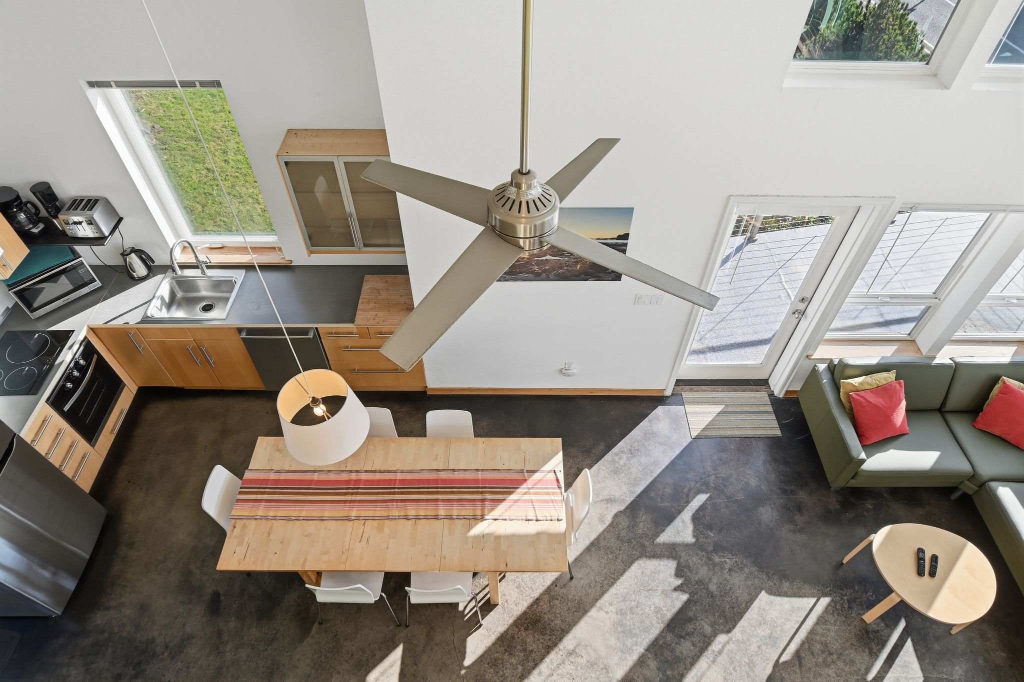 Overhead view of an open-concept dining and kitchen area with dark polished concrete floors. A large wooden dining table with white chairs sits under a modern ceiling fan, adjacent to a light wood kitchen with stainless steel appliances.