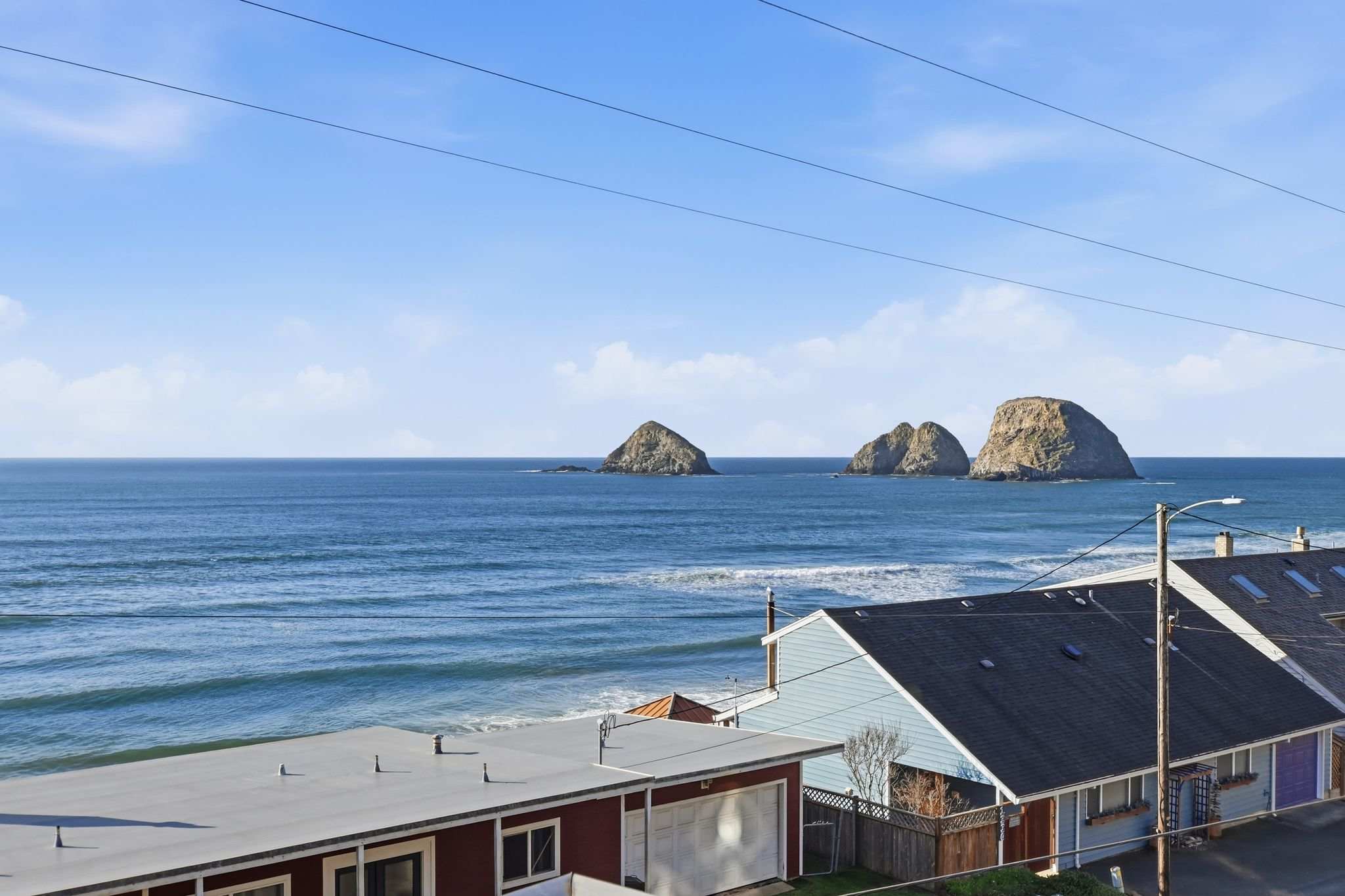 A wide shot of the ocean with prominent rock formations in the distance, showing waves breaking on the shore, with rooftops of nearby houses in the foreground under a bright blue sky.