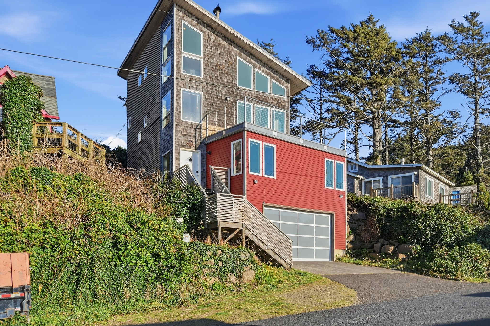 The multi-level house features a distinctive red lower section with large windows and a garage door, topped by a shingle-sided upper structure with numerous windows and a sloped roof. Lush green foliage and tall trees surround the property, which is accessed by a paved driveway and wooden stairs.