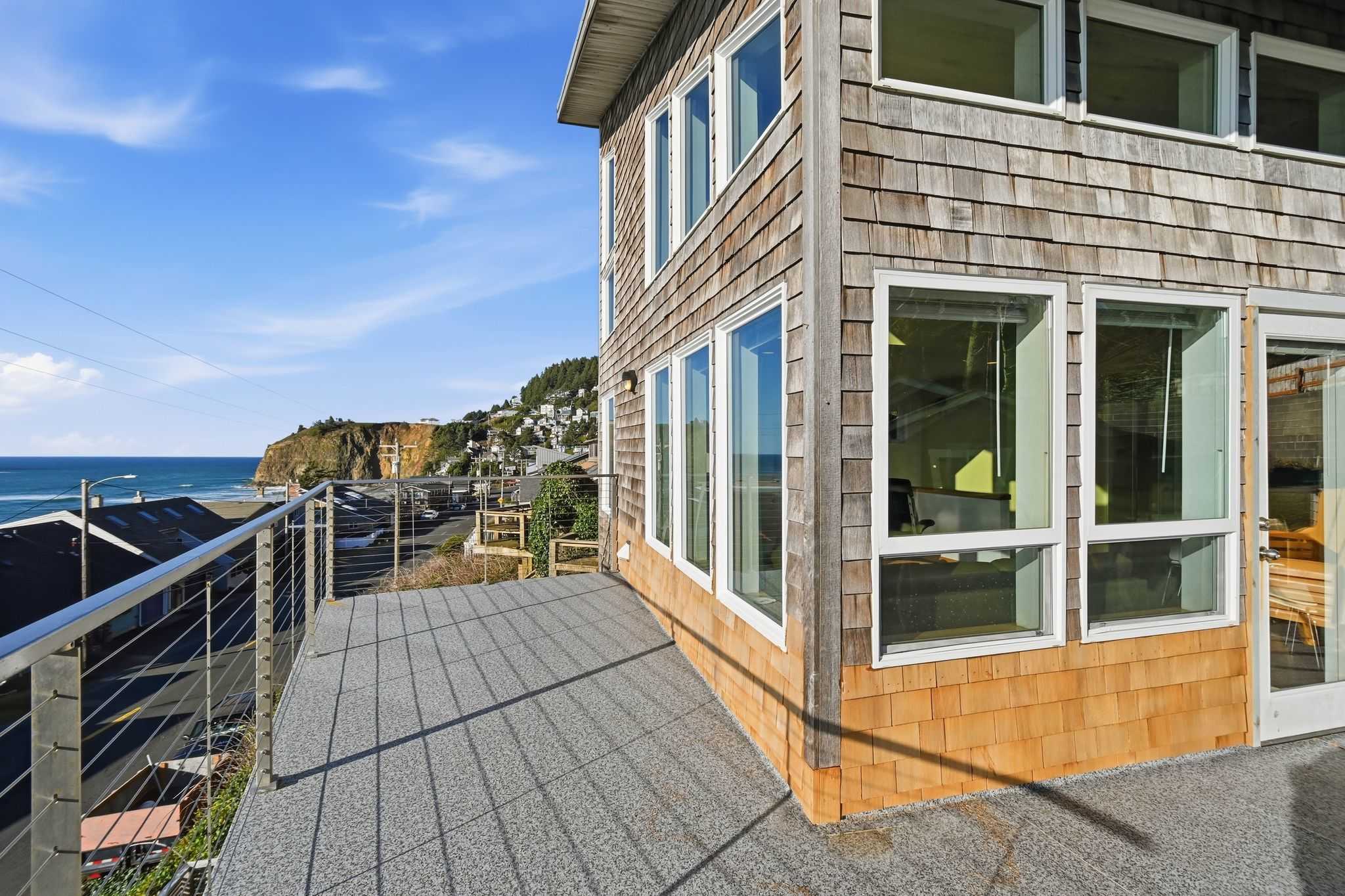 A modern balcony with metal railings offers a panoramic view of the ocean and the coastal town, featuring a house with shingle siding and large windows. The deck floor is textured gray, reflecting the bright blue sky.