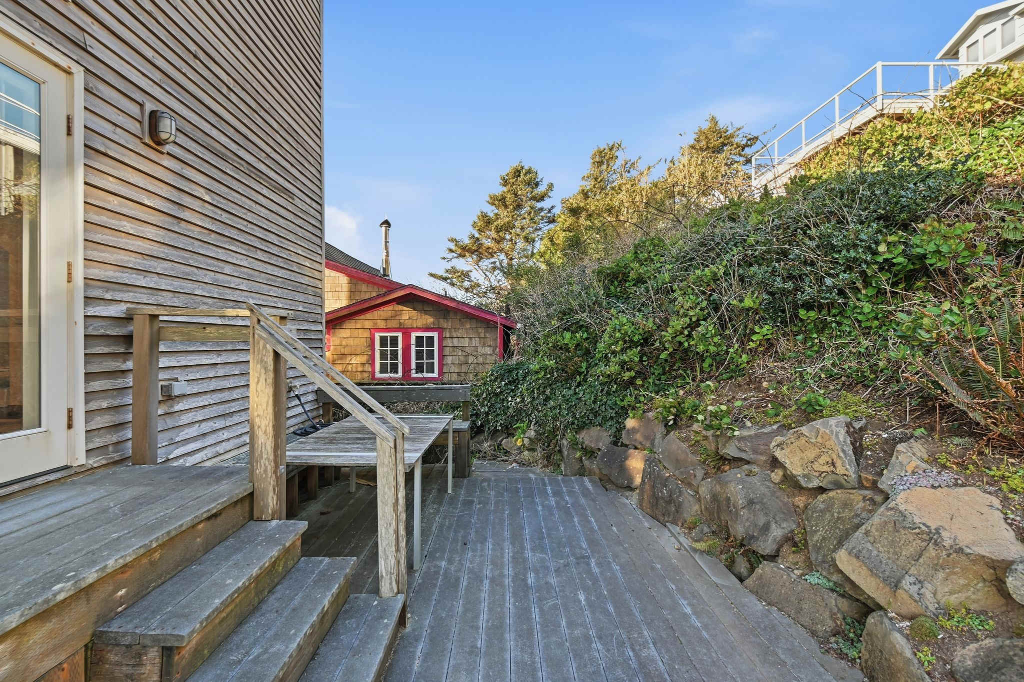 A weathered wooden deck extends towards a natural rock wall and lush green foliage, with a small red-sided shed visible in the background. Wooden steps lead up to the house entrance, which is clad in horizontal wooden siding.