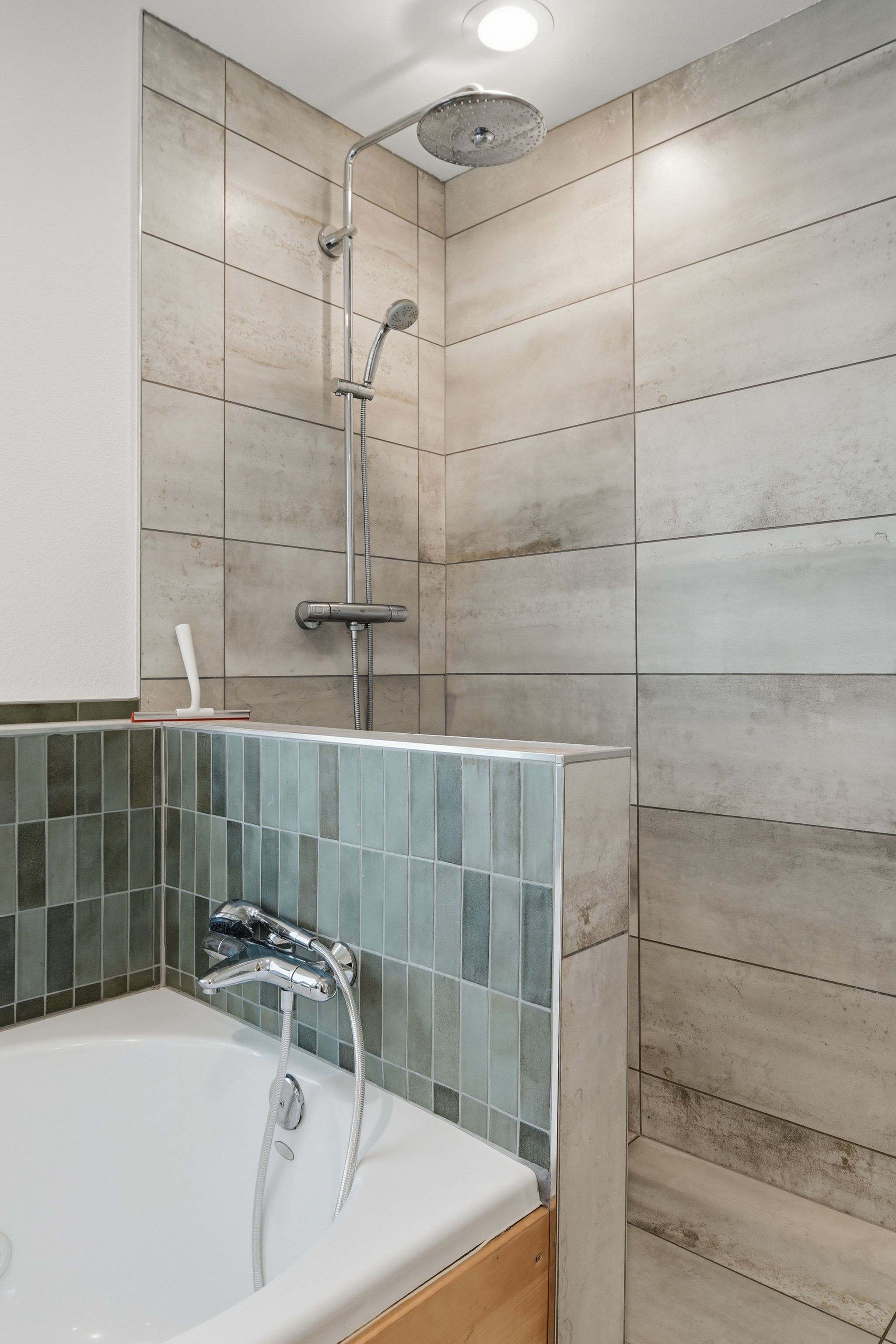 A detailed view of the shower area in a bathroom, featuring a rain showerhead and a handheld shower. The walls are tiled in light grey and green, with a partial divider separating it from the tub.