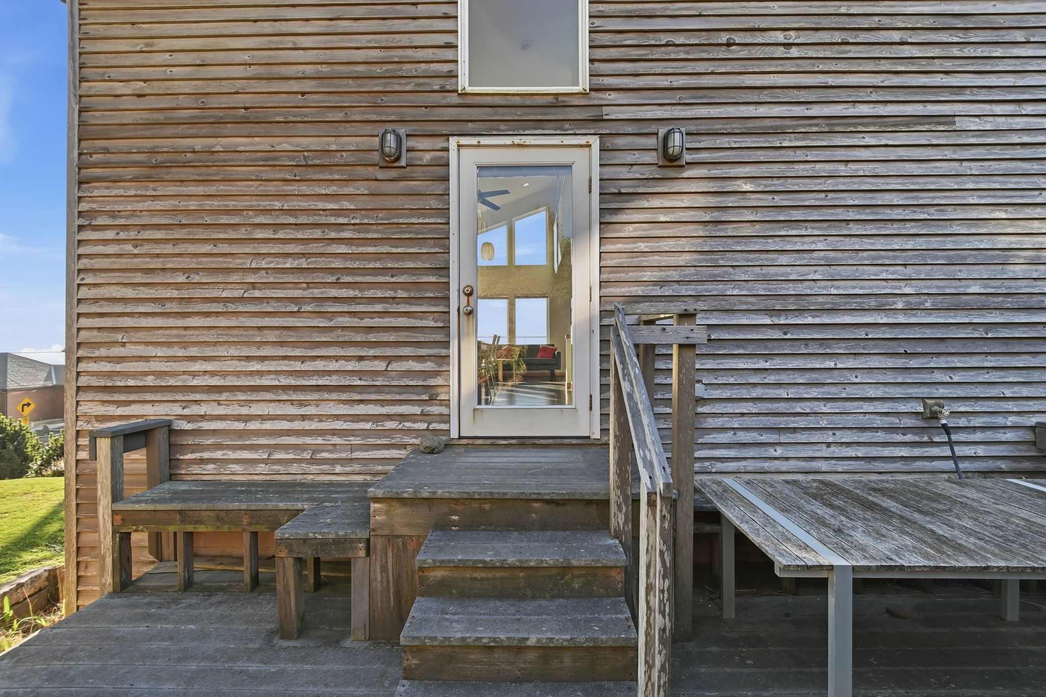 A rustic wooden deck features steps leading up to a glass-paned door, flanked by two outdoor wall sconces. The house exterior is clad in weathered wooden planks, with a small wooden bench built into the deck.