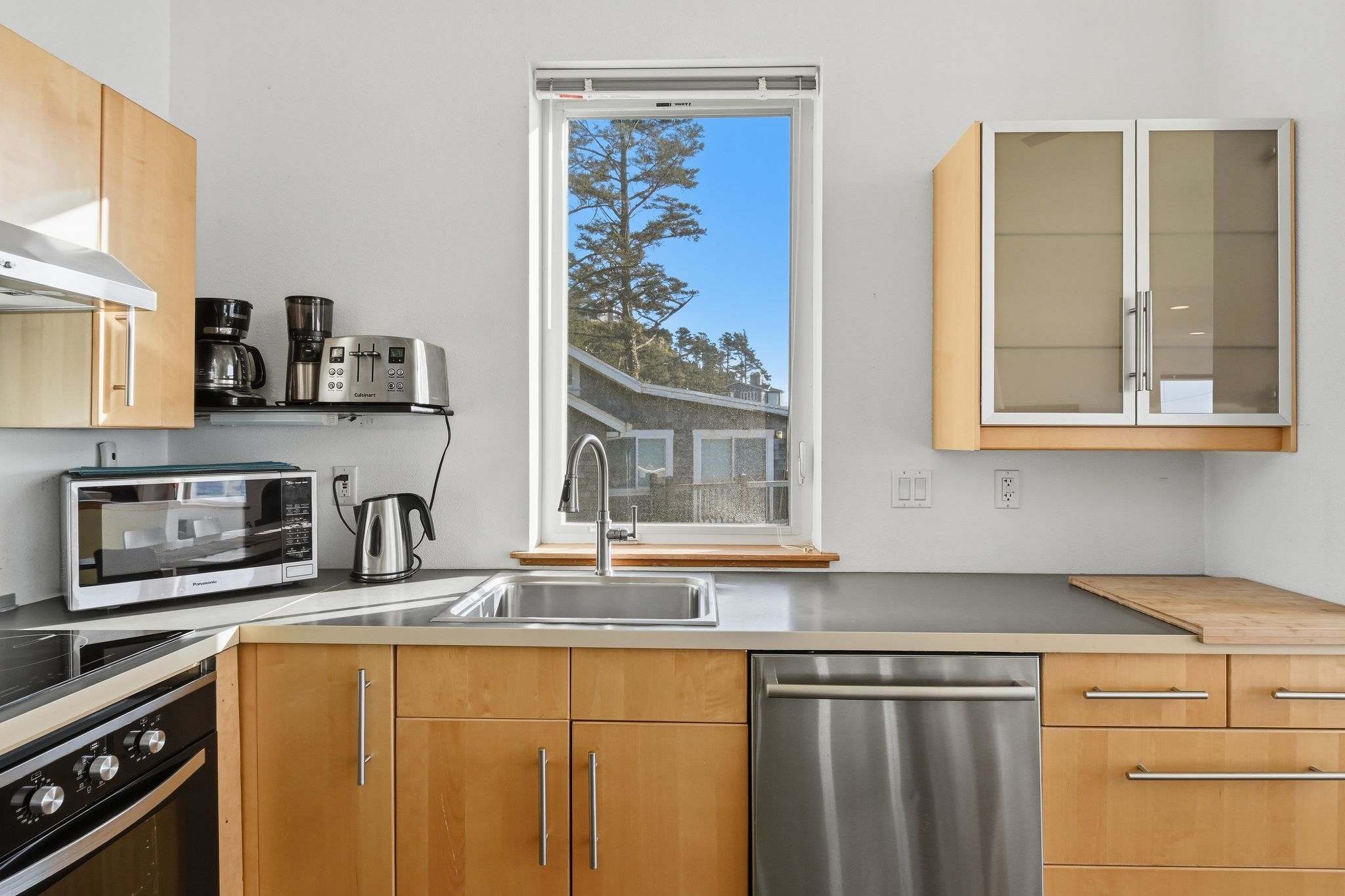 Detailed view of the kitchen counter with a stainless steel sink and modern faucet, under a tall window. Appliances include a coffee maker, toaster, and microwave, set against light wood cabinetry.