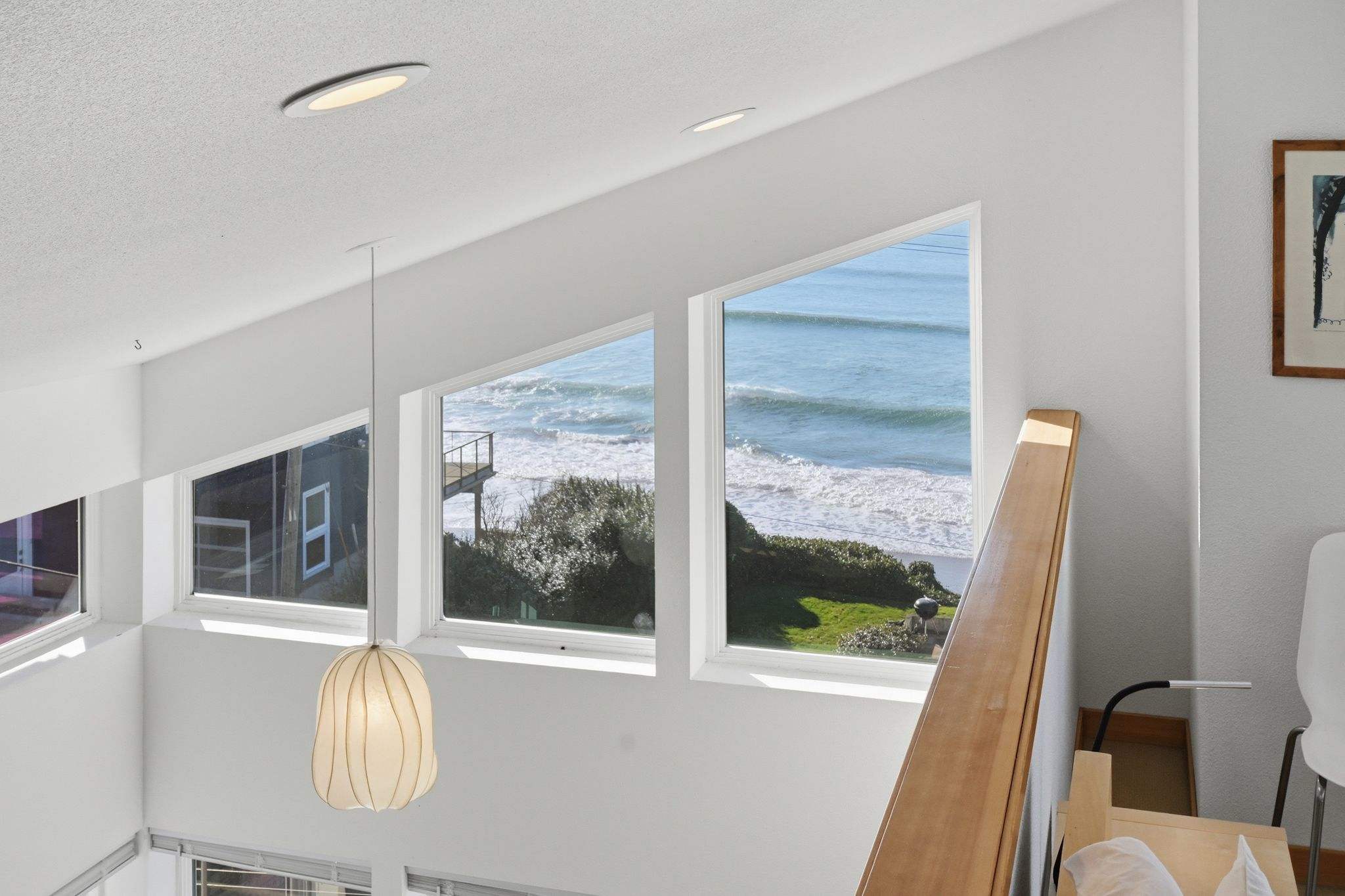 A bright staircase landing features multiple large, angled windows providing a stunning view of the ocean and breaking waves, with a modern pendant light hanging from the high ceiling.