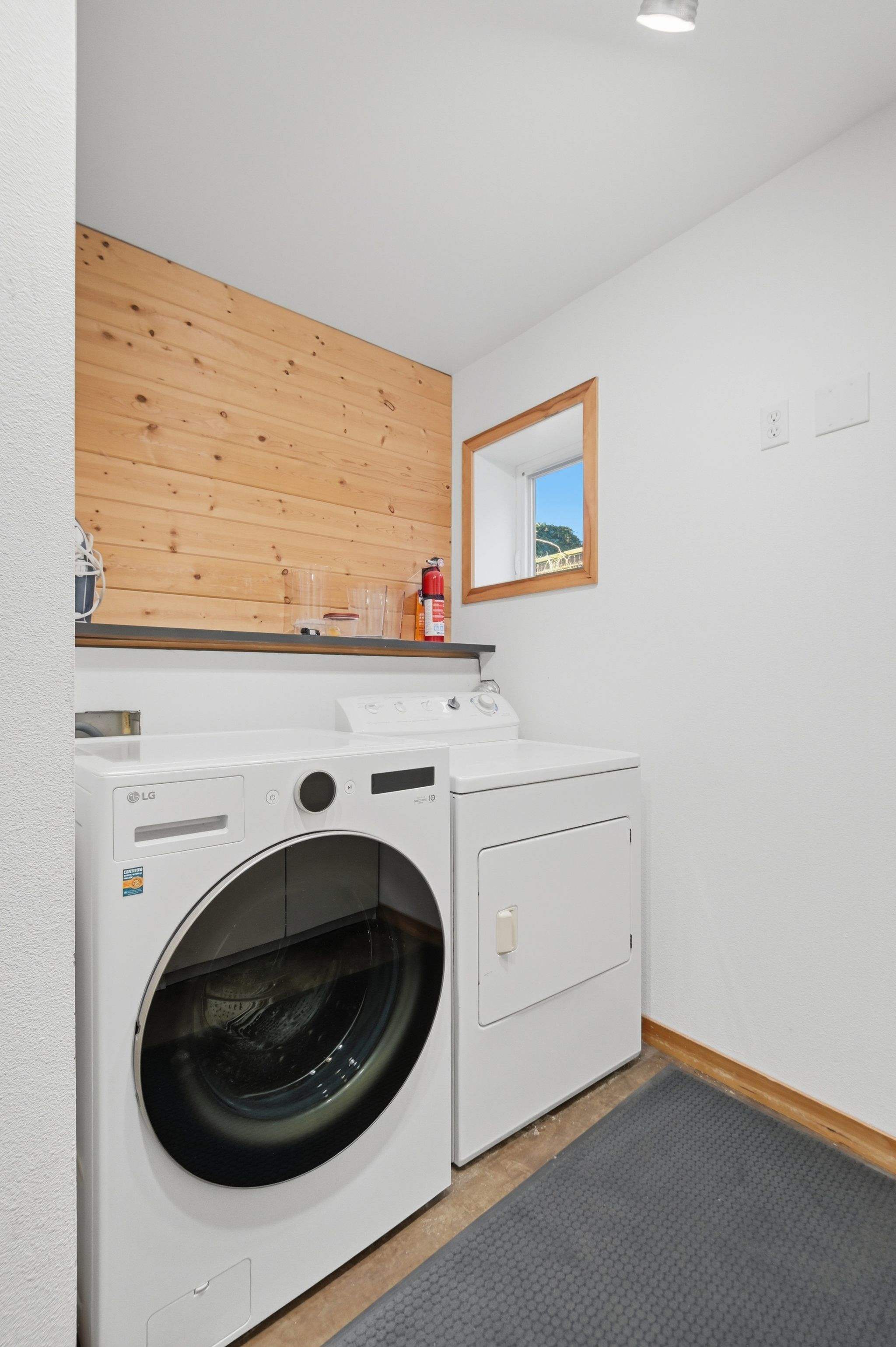 A functional laundry room contains a white LG front-load washing machine and a matching dryer, set against white walls with a natural wood accent wall and a small window.