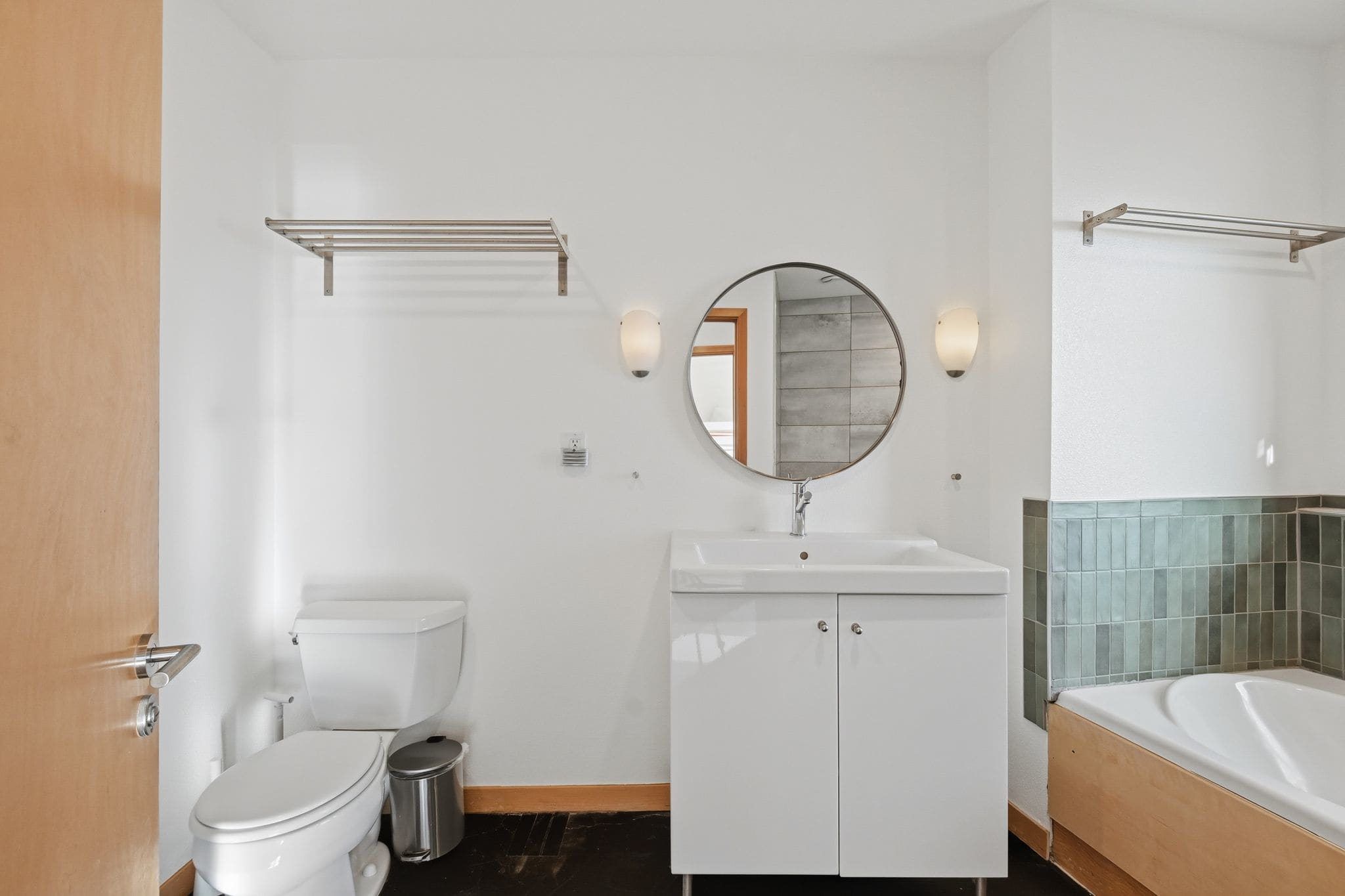 A full view of a modern bathroom with a white vanity, round mirror, toilet, and a jetted bathtub with an integrated shower. The walls feature green and light grey tiling, and dark flooring.