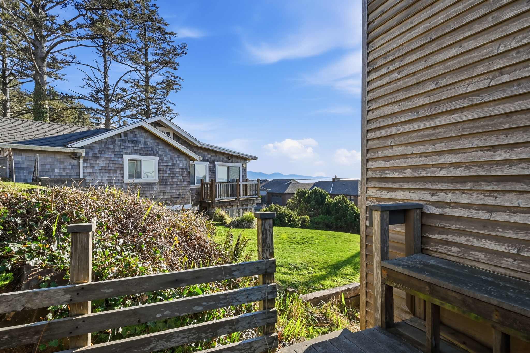 From a wooden deck with a built-in bench, a view opens to a well-maintained grassy lawn and distant ocean horizons. Another shingle-sided house with a deck is visible across the lawn, framed by tall trees and a bright blue sky.