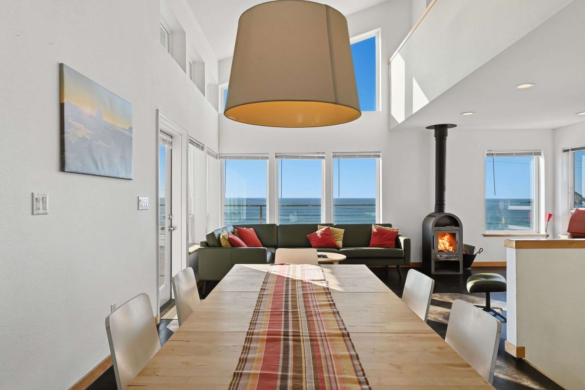 Dining area with a long wooden table and white chairs, illuminated by a large drum pendant light. Beyond, the living room features a green sofa and expansive windows showcasing direct ocean views.