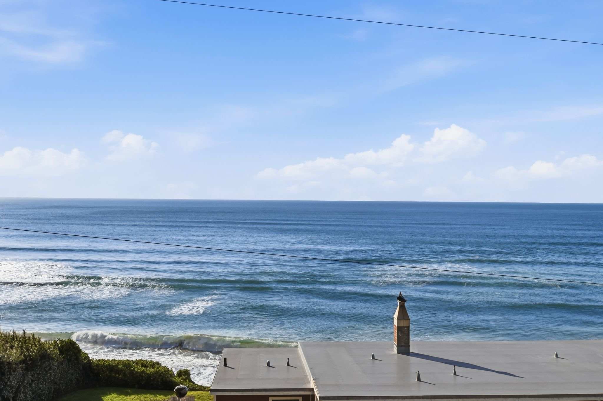 An expansive view of the deep blue ocean with gentle waves, seen over the flat roof of a neighboring building, under a clear sky with scattered clouds, emphasizing the vast coastal landscape.