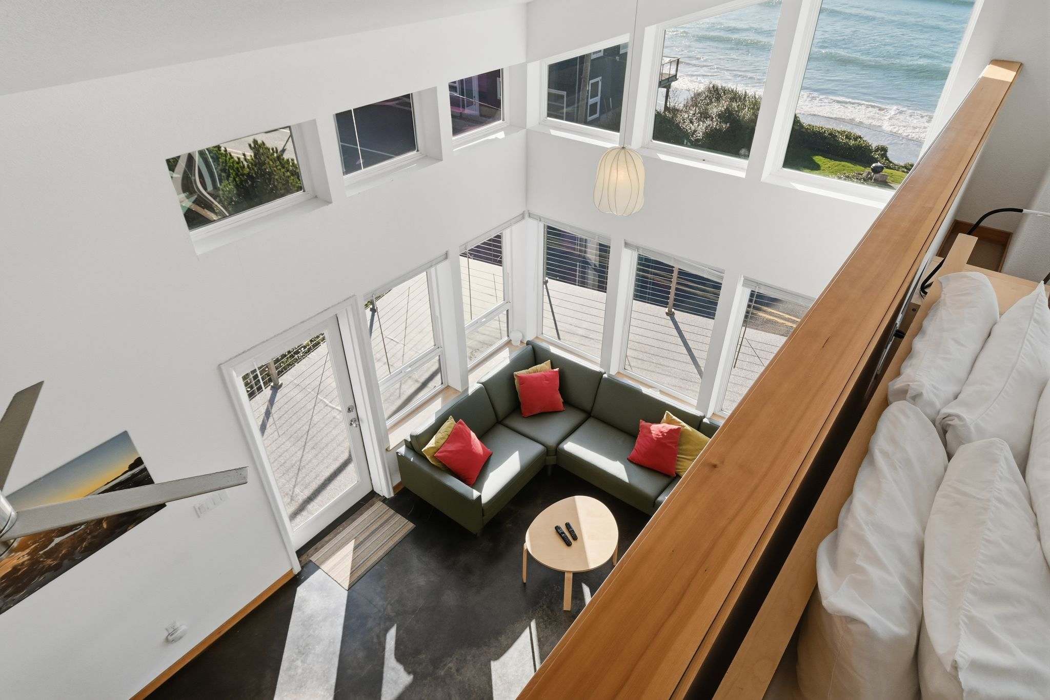 Aerial view of a bright living room featuring a green sectional sofa with red and yellow throw pillows. Large windows offer panoramic ocean views, and a small round coffee table sits on dark flooring.