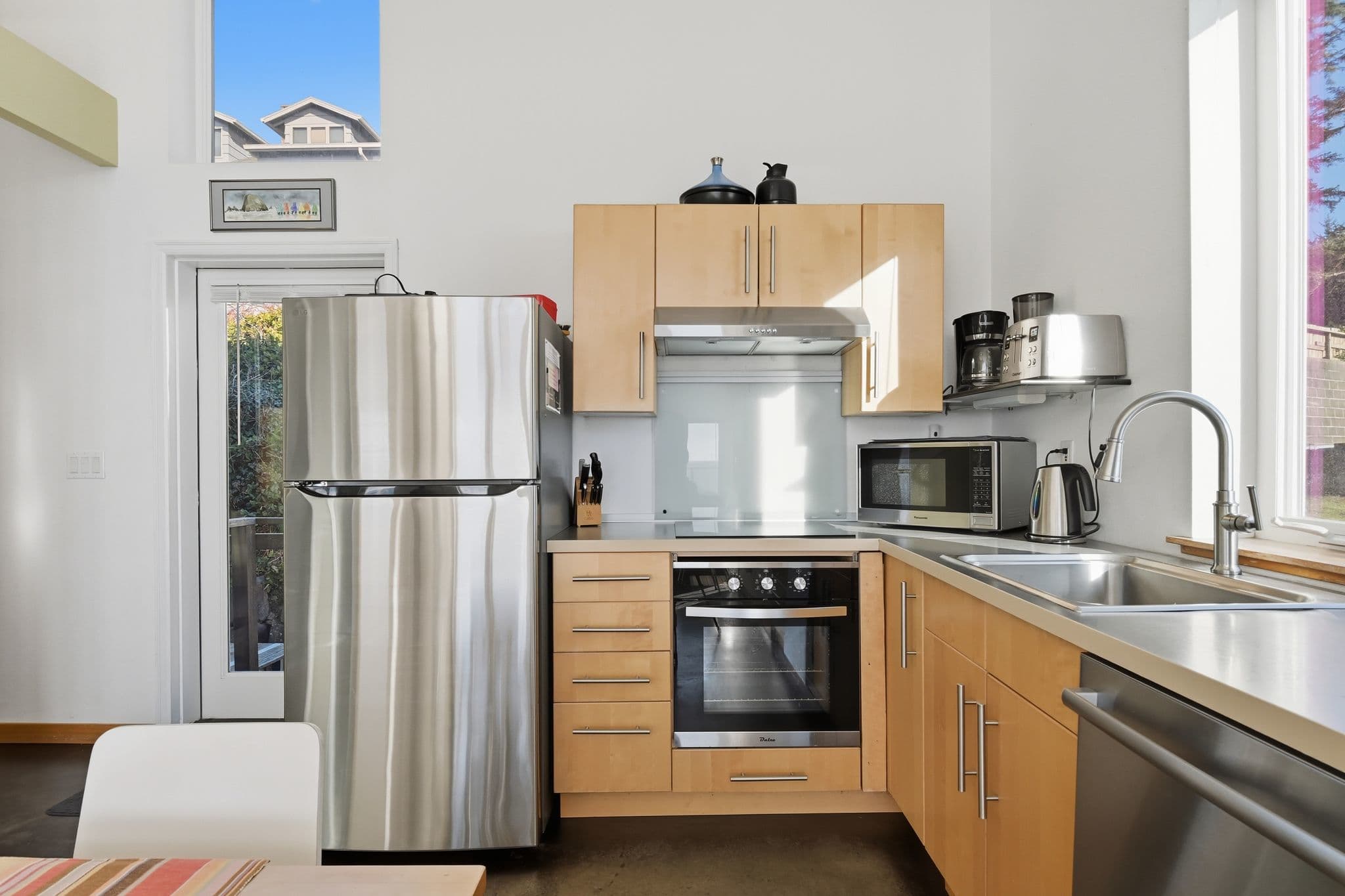 Modern kitchen with light wood cabinetry and stainless steel appliances, including a refrigerator and built-in oven. A window above the sink offers a view of the exterior, and a door leads outside.