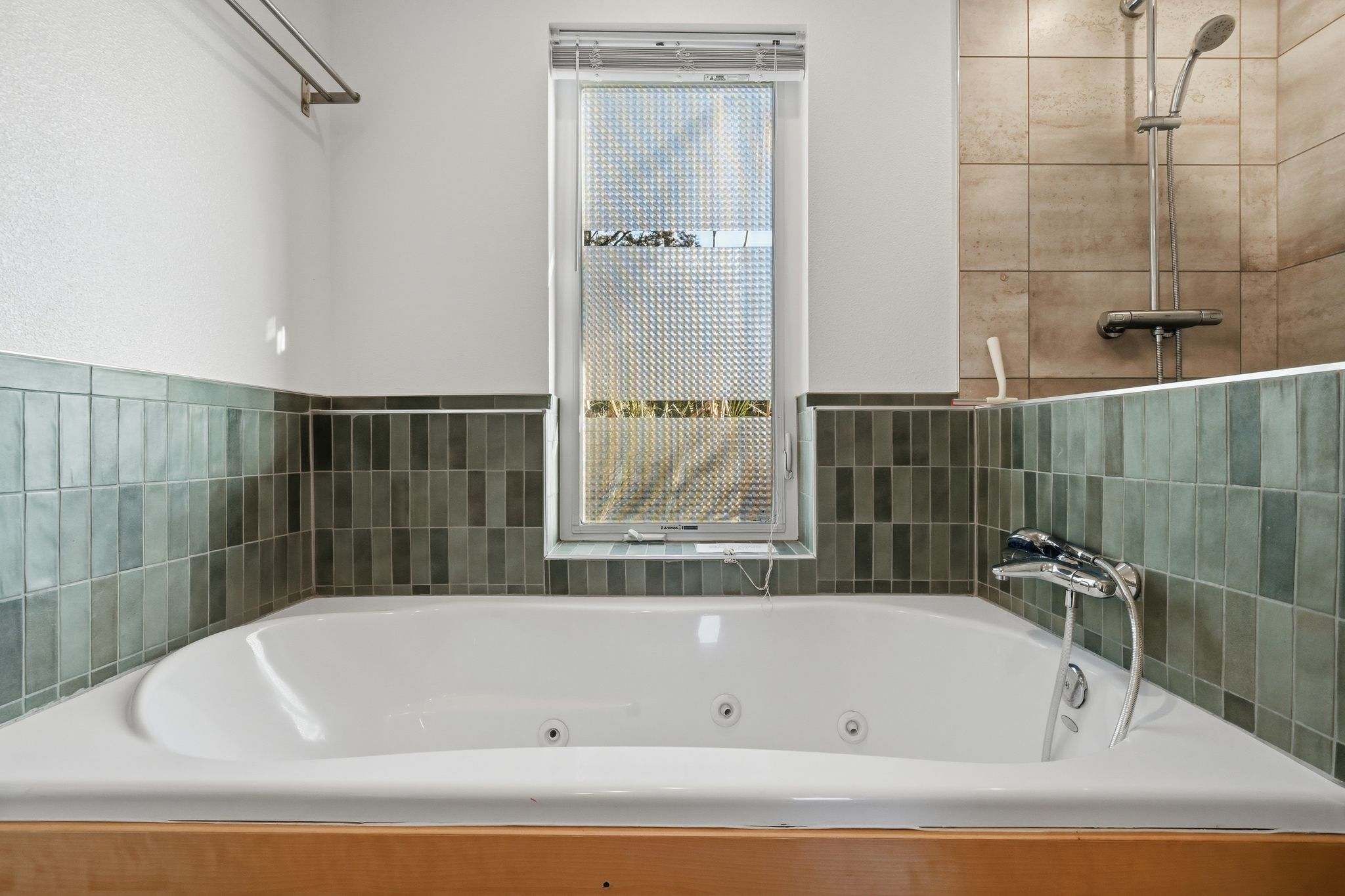 A close-up of the white jetted bathtub with a wooden surround, showing the jets and the shower/faucet fixtures. A window with privacy blinds is positioned above the tub.