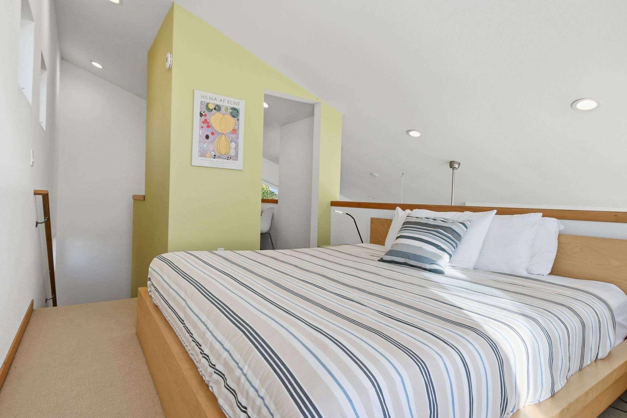 A bright bedroom featuring a light wood bed frame with striped bedding, set against a light yellow accent wall. A modern art print is visible, and the room has recessed lighting and a sloped ceiling.