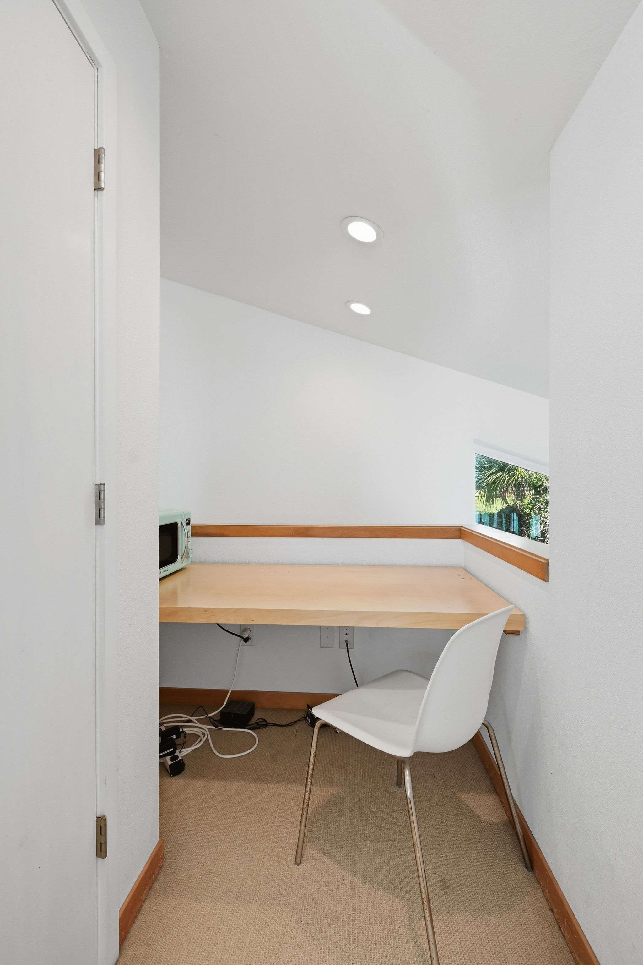 A compact office nook features a light wood desk, a white modern chair, and a retro mint green microwave, with a small window offering a glimpse of greenery. The space has white walls and recessed lighting.