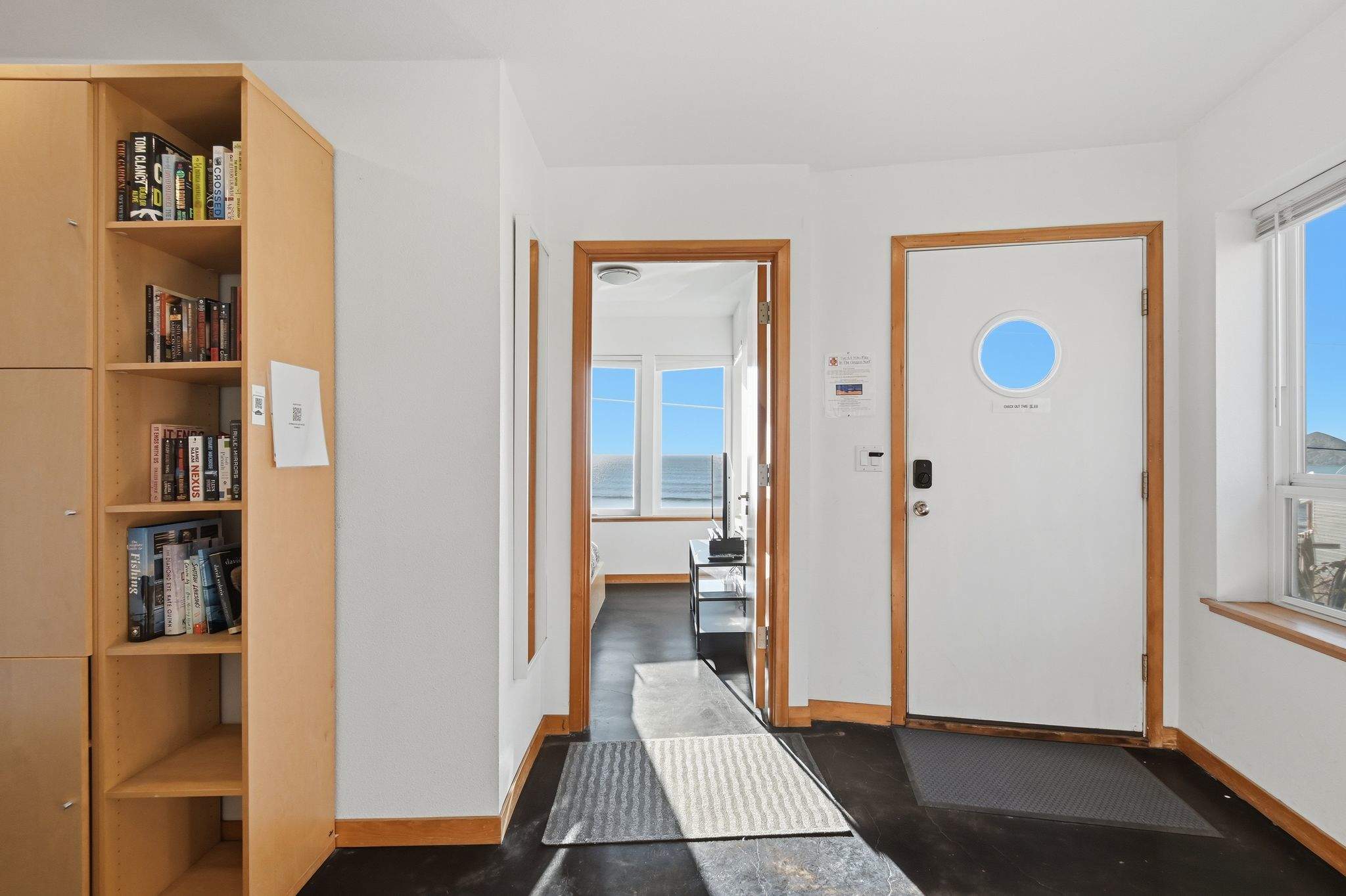 Bright foyer with dark flooring, a tall wooden bookshelf filled with books, and a white door featuring a porthole window. A doorway leads to a room with expansive ocean views.