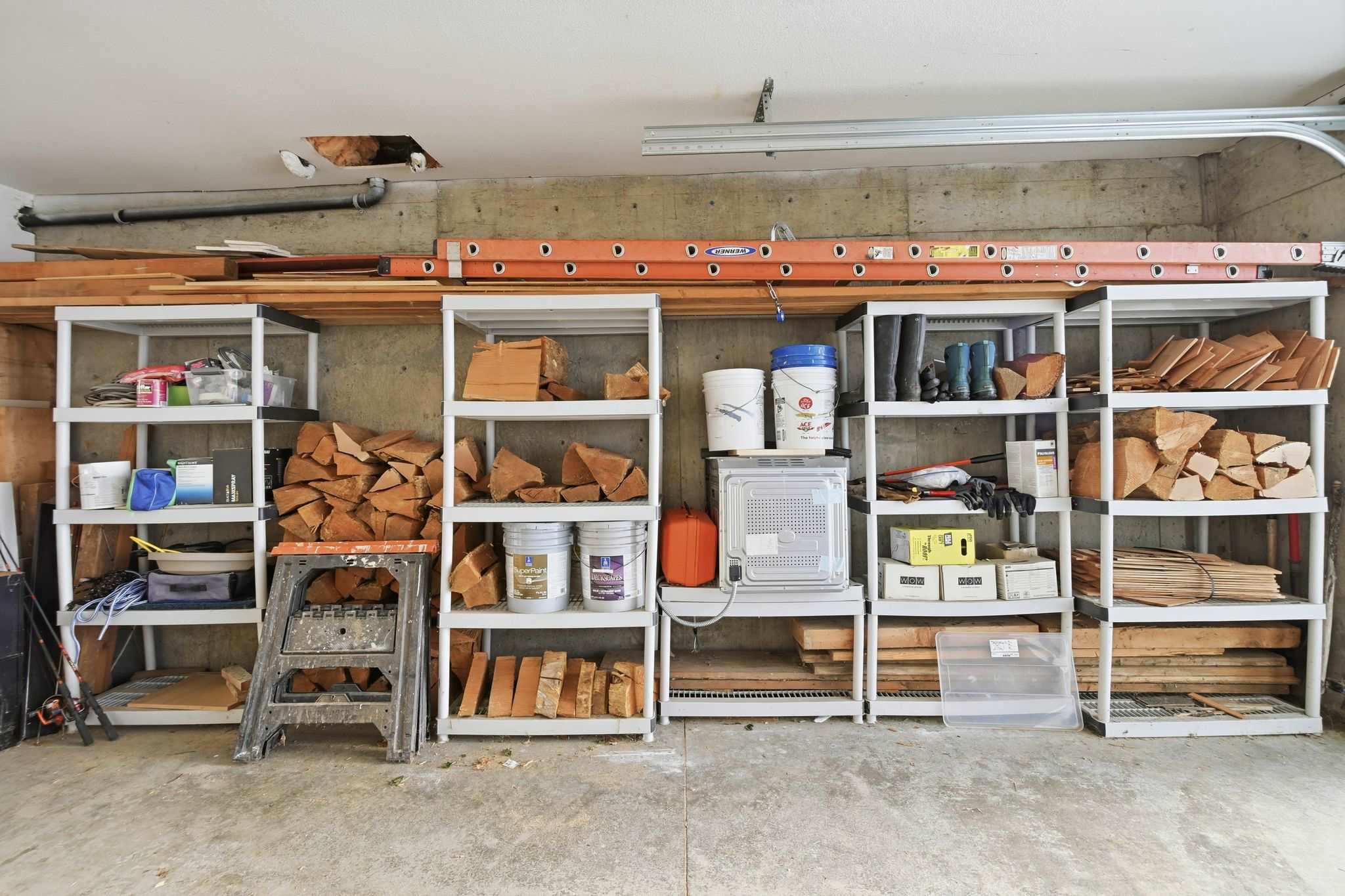 A garage or storage area features multiple white plastic shelving units filled with neatly stacked firewood, various buckets, and tools, set against a concrete wall.