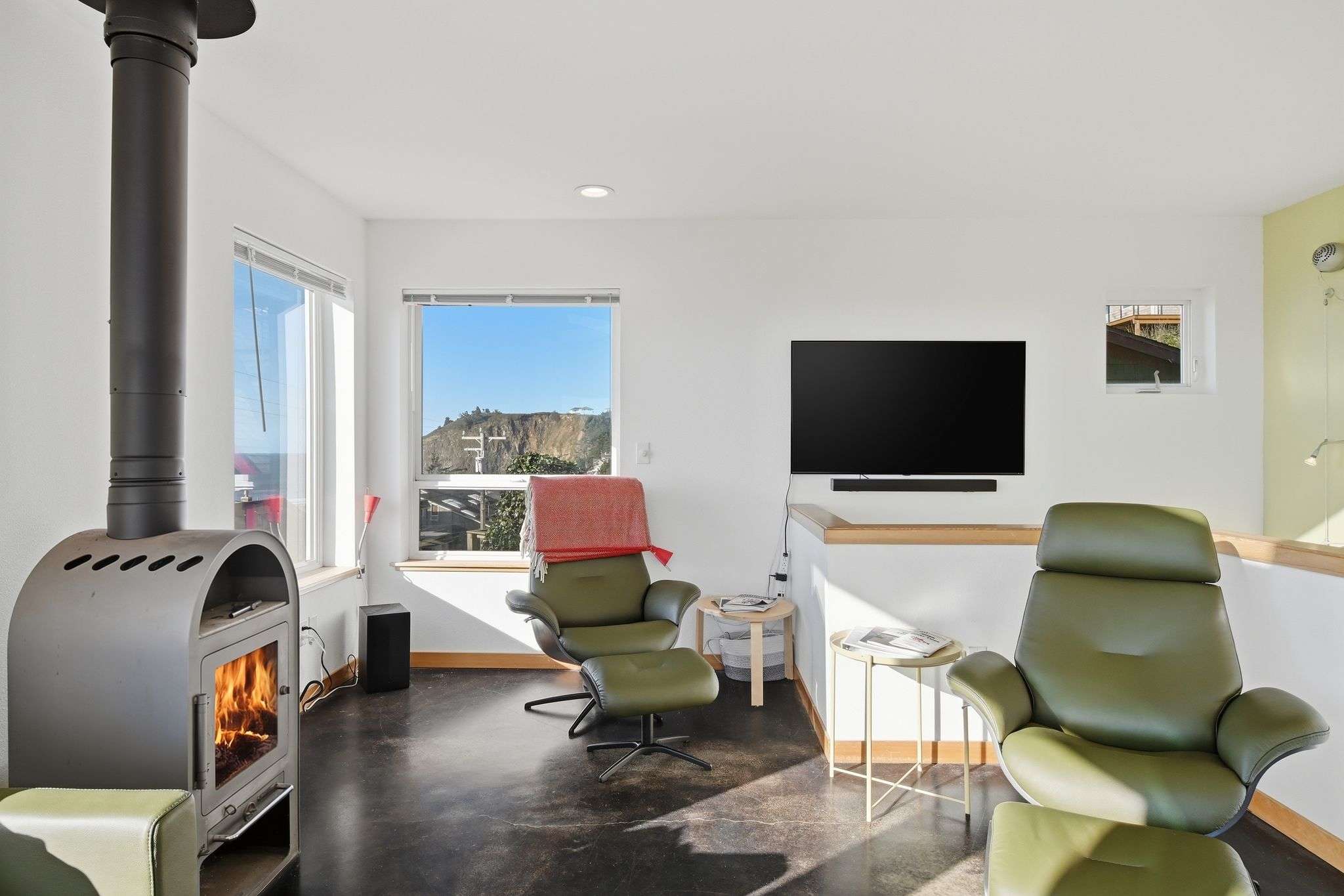 A cozy living room features a modern wood-burning stove, two olive green leather armchairs with ottomans, and large windows offering views of the surrounding neighborhood and distant hills.