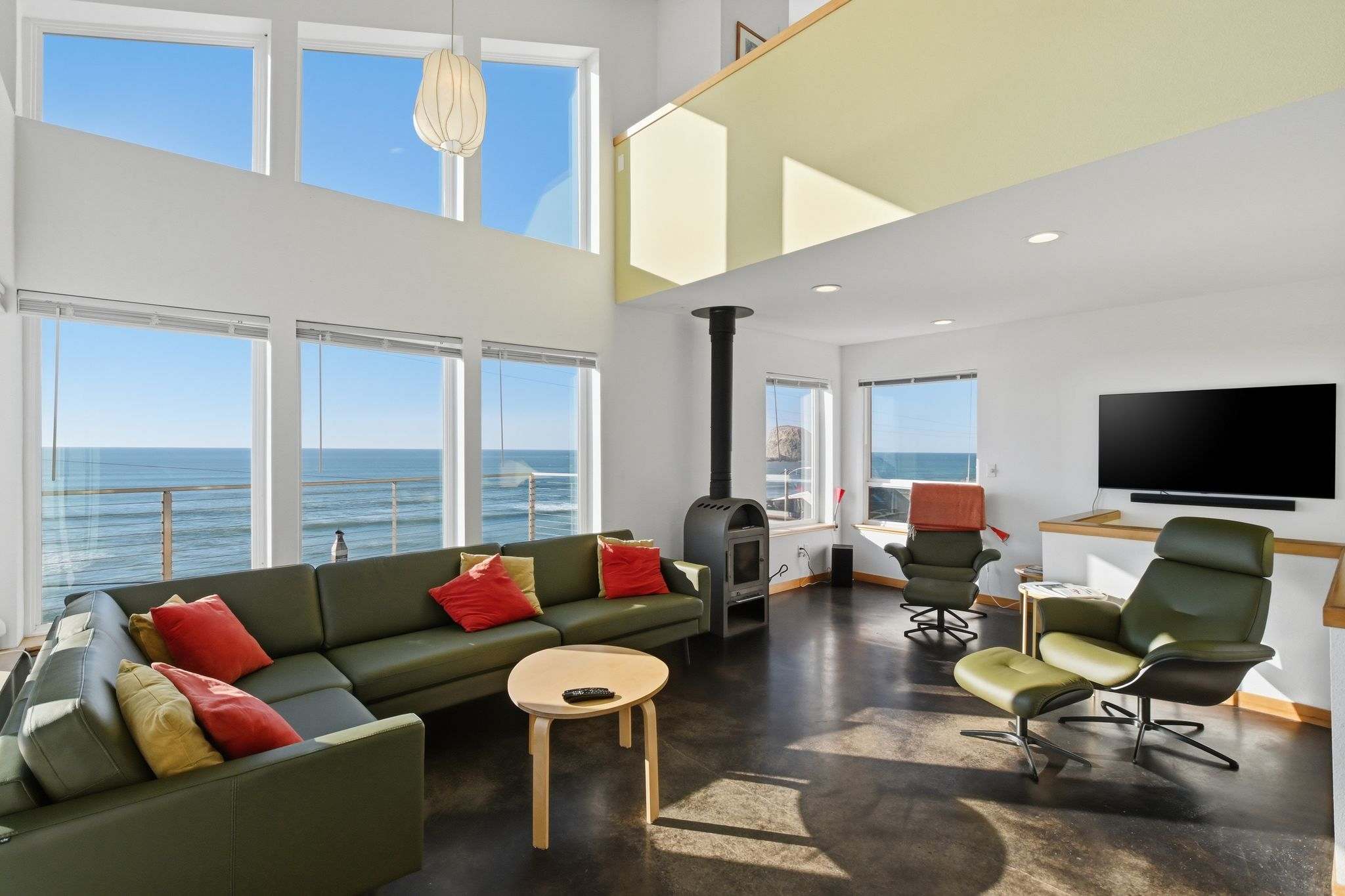 A spacious living room boasts floor-to-ceiling windows providing expansive ocean views, furnished with a large olive green sectional sofa, a modern wood-burning stove, and polished concrete floors.