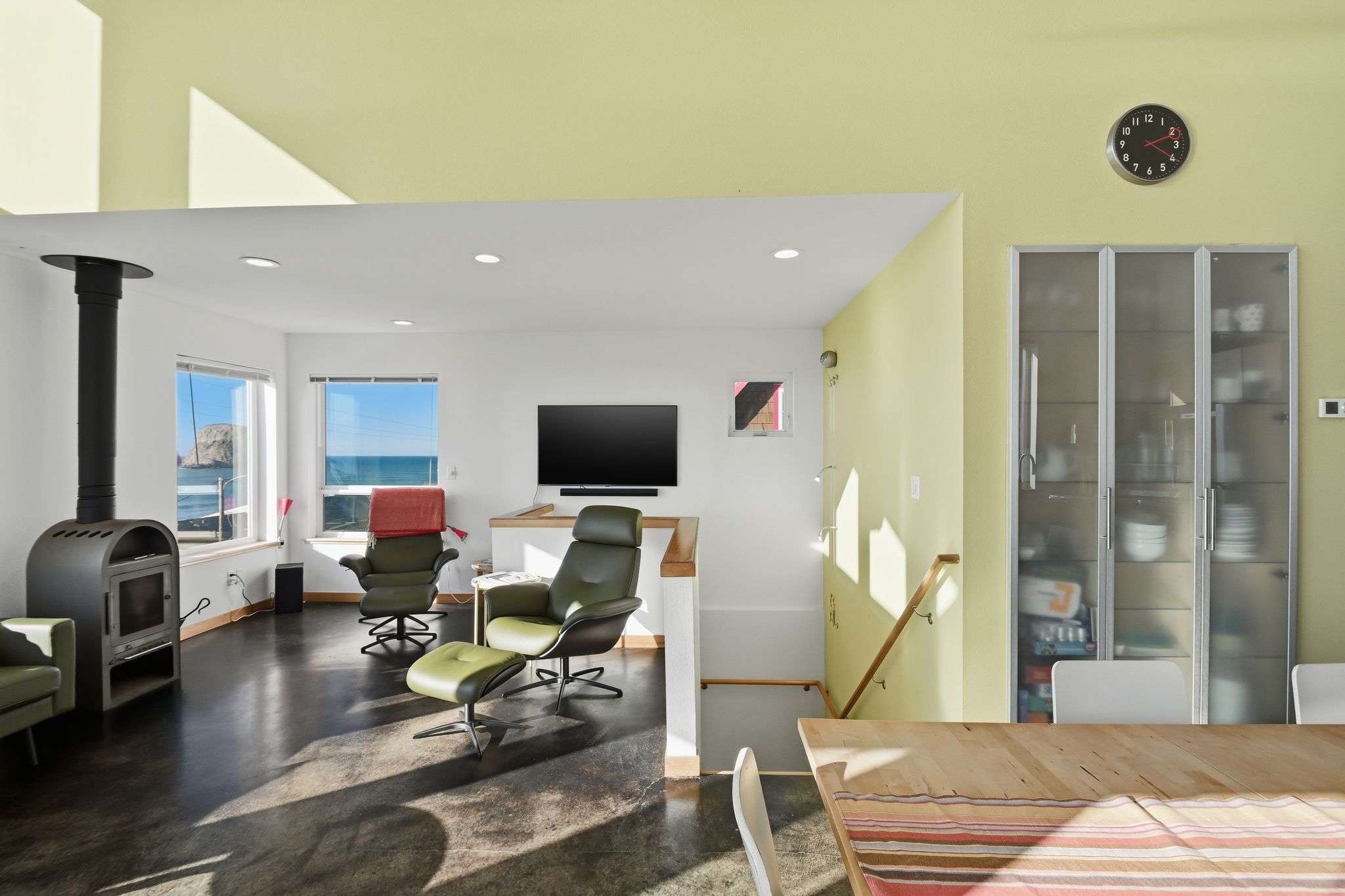 Spacious living room with dark flooring, featuring a modern wood-burning stove and two green leather armchairs with ottomans. A wall-mounted TV is visible, and large windows provide stunning ocean views.