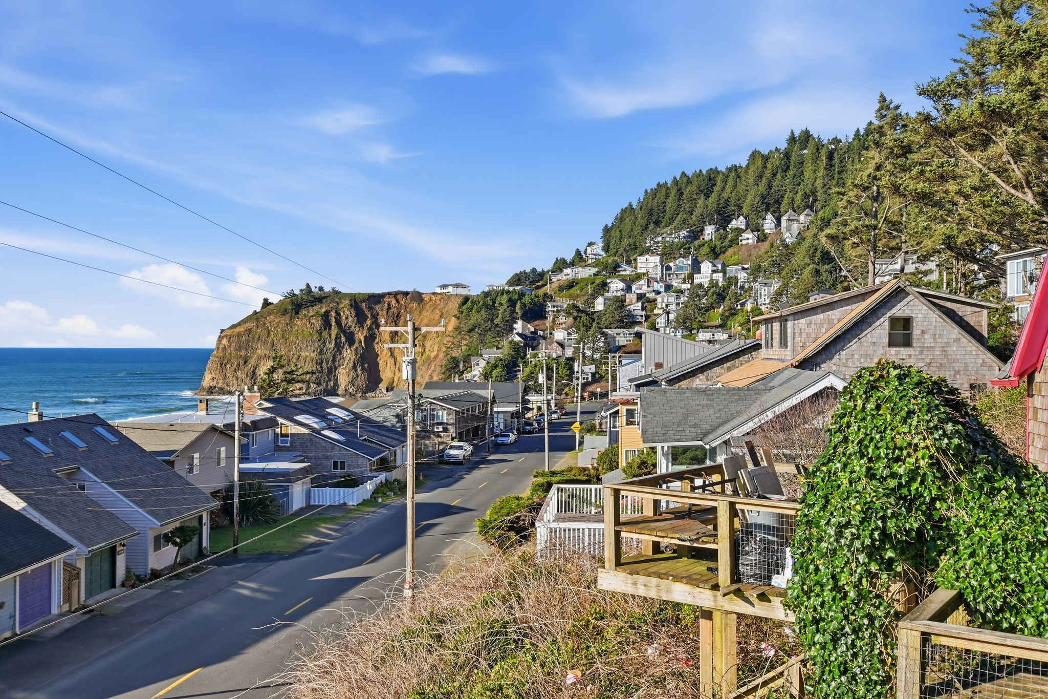 A scenic aerial view captures a coastal town nestled on a hillside, with a winding road leading towards the ocean and a dramatic cliff face. Numerous houses with various rooflines are visible, surrounded by lush green trees under a bright blue sky.