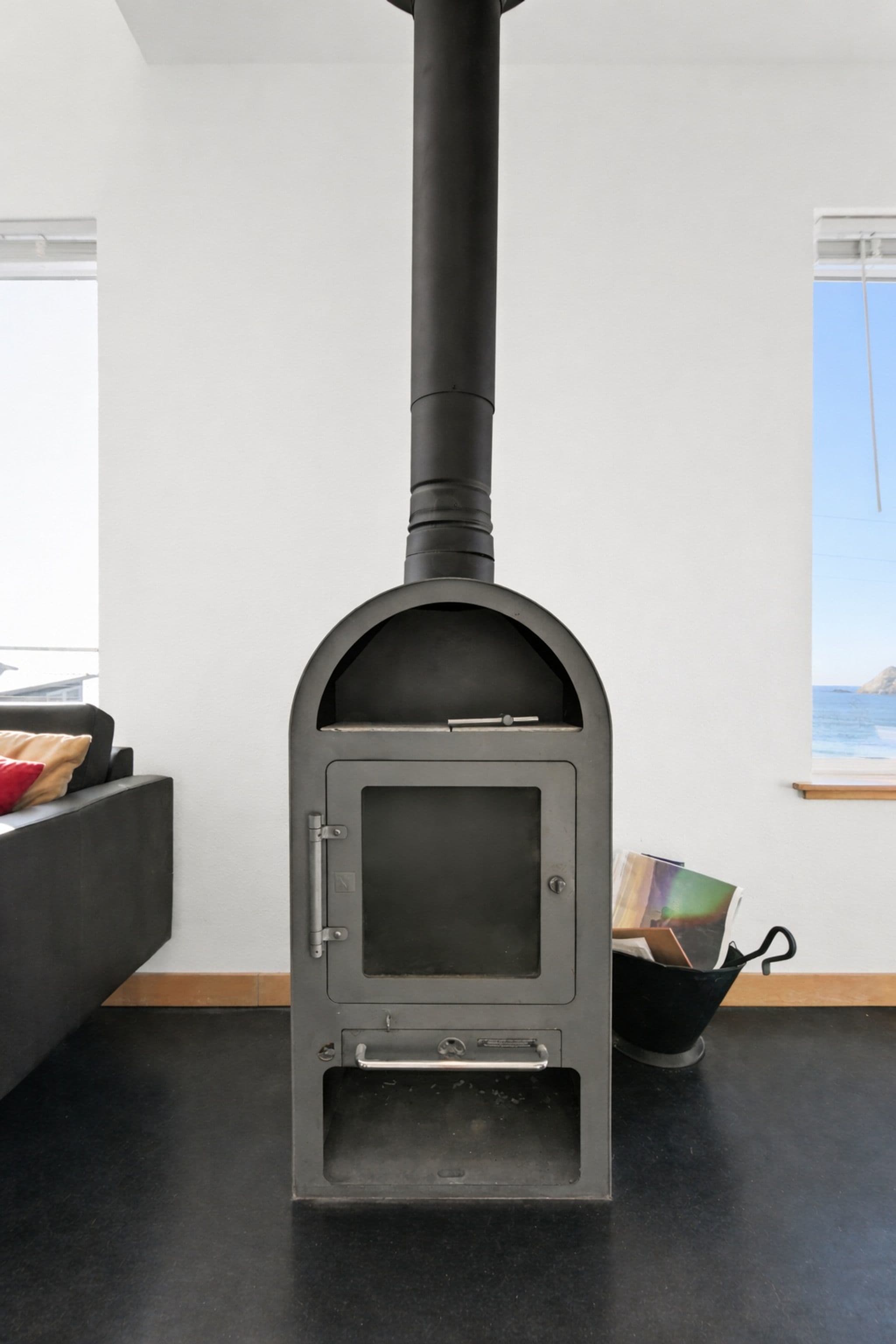 A modern black wood-burning stove with a tall flue stands on a dark floor in a bright room with white walls and windows offering a glimpse of the ocean.