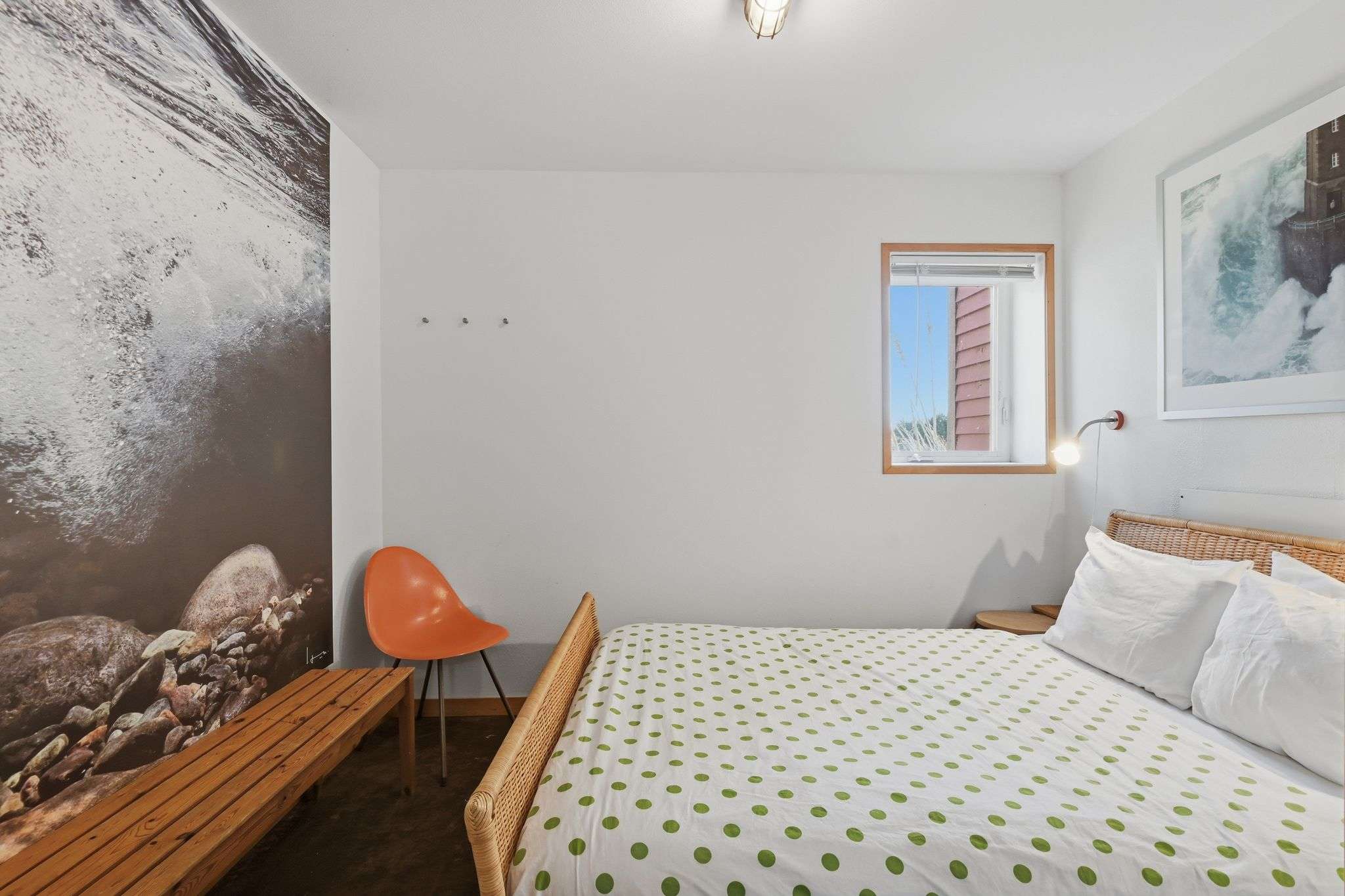 A cozy bedroom featuring a bed with a woven headboard and green polka-dot bedding. A large mural covers one wall, and a small window with a wooden frame provides natural light.