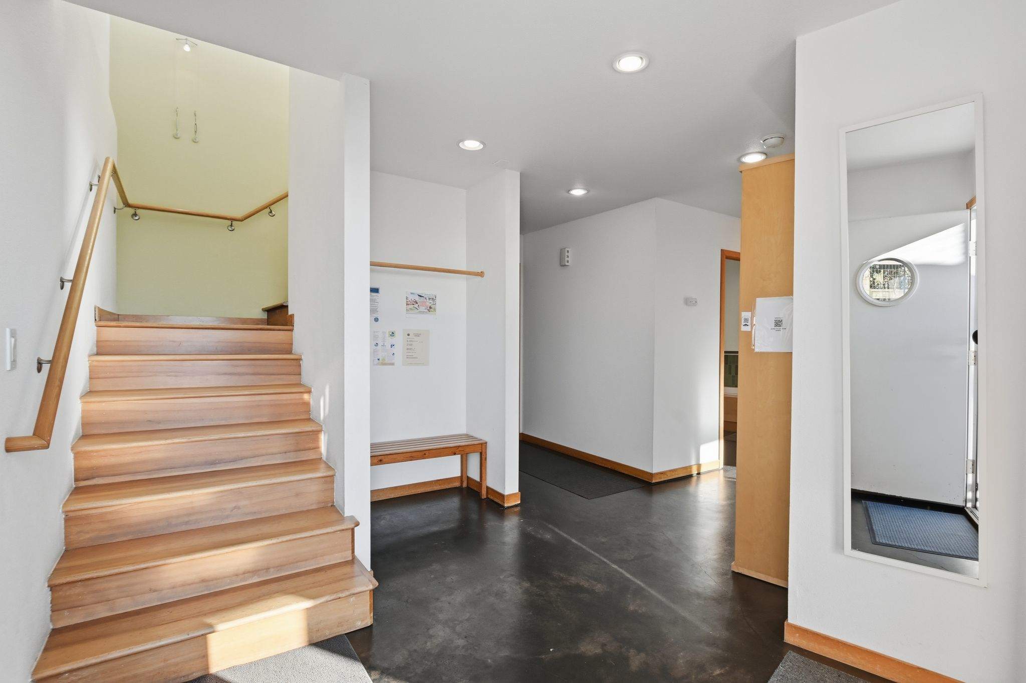 A bright entryway features a wooden staircase leading upstairs, dark flooring, white walls, and a small wooden bench with a coat rack, along with a full-length mirror.