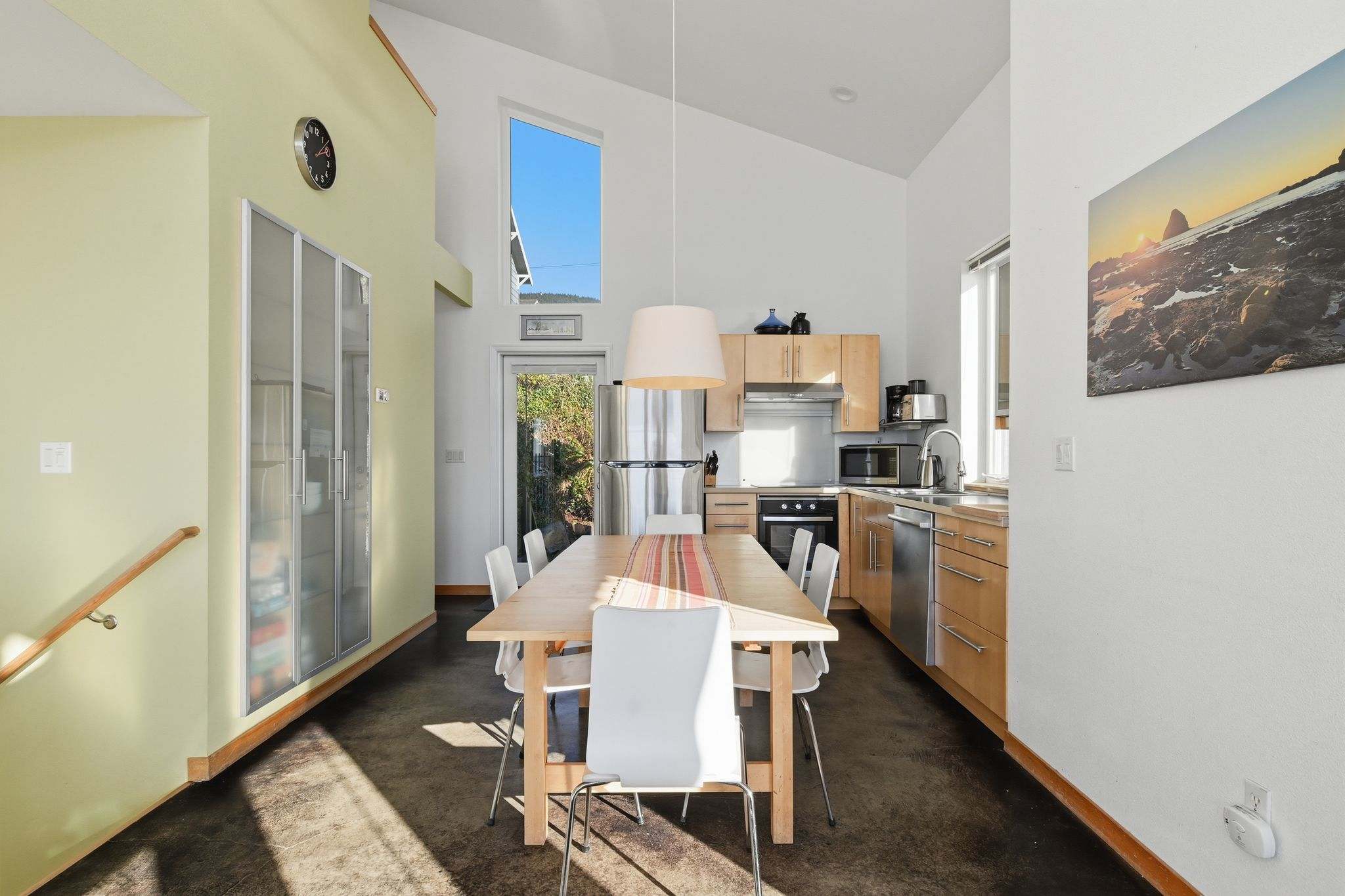 Open-concept dining and kitchen area with high ceilings and dark flooring. A wooden dining table is central, with the kitchen visible to the right and a tall, frosted-glass cabinet to the left.