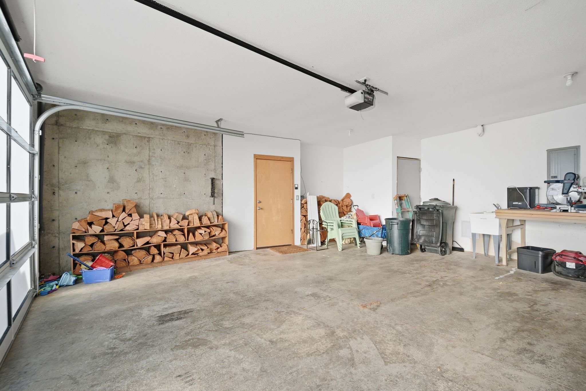 A spacious garage with a concrete floor features a large stack of firewood neatly organized against a concrete wall. Various outdoor chairs, bins, and tools are stored along the white-painted walls, with a garage door visible on the left.