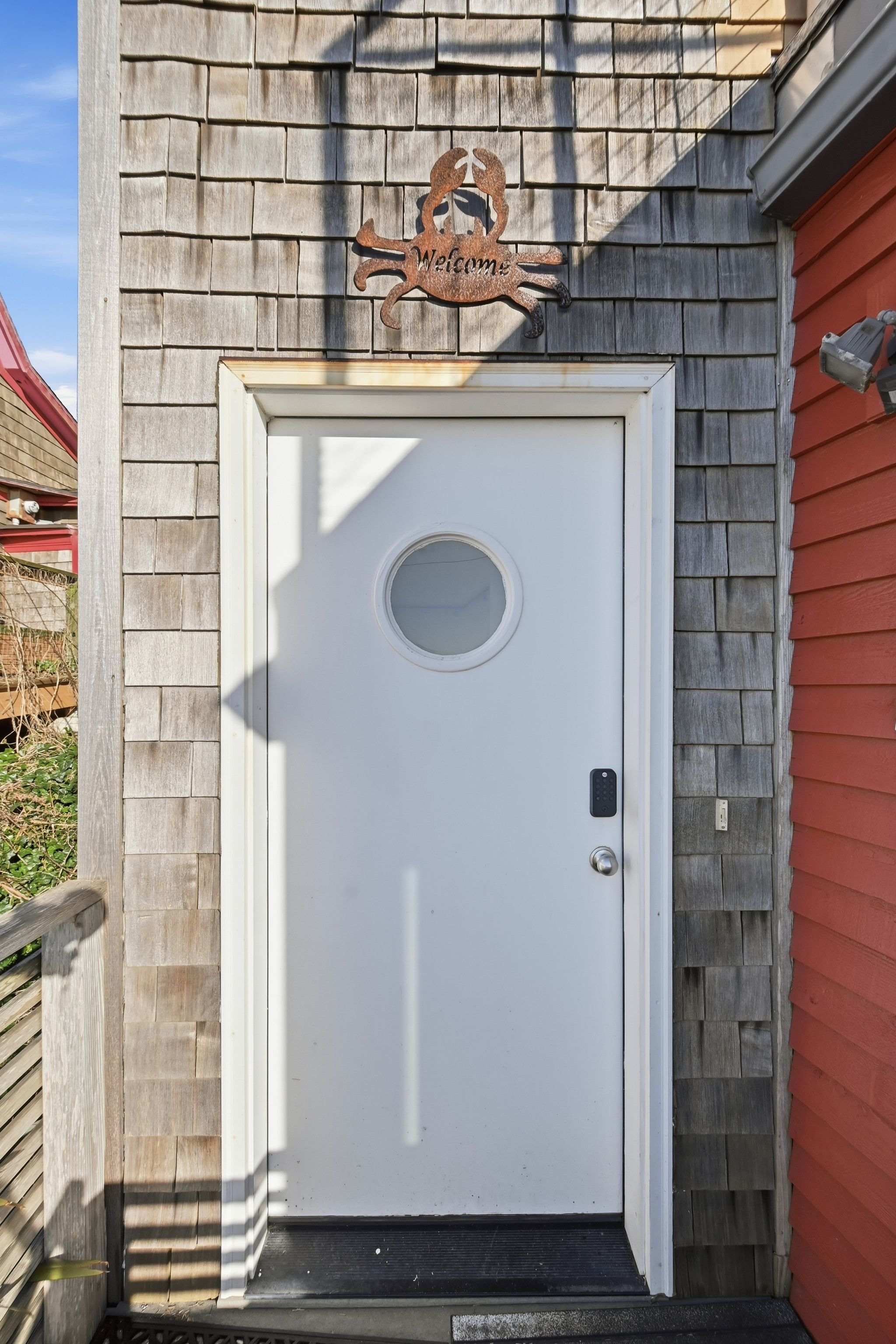 A white exterior door with a round porthole window is set into a shingle-sided wall, adorned with a rustic metal crab "Welcome" sign. The door features a modern keyless entry system and a simple doorknob.